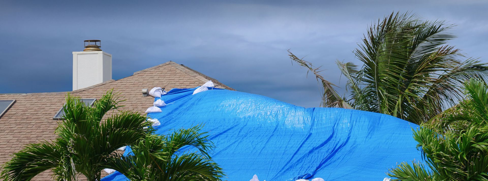 A blue tarp covers a damaged roof, trees in the foreground, white chimney, stormy sky.