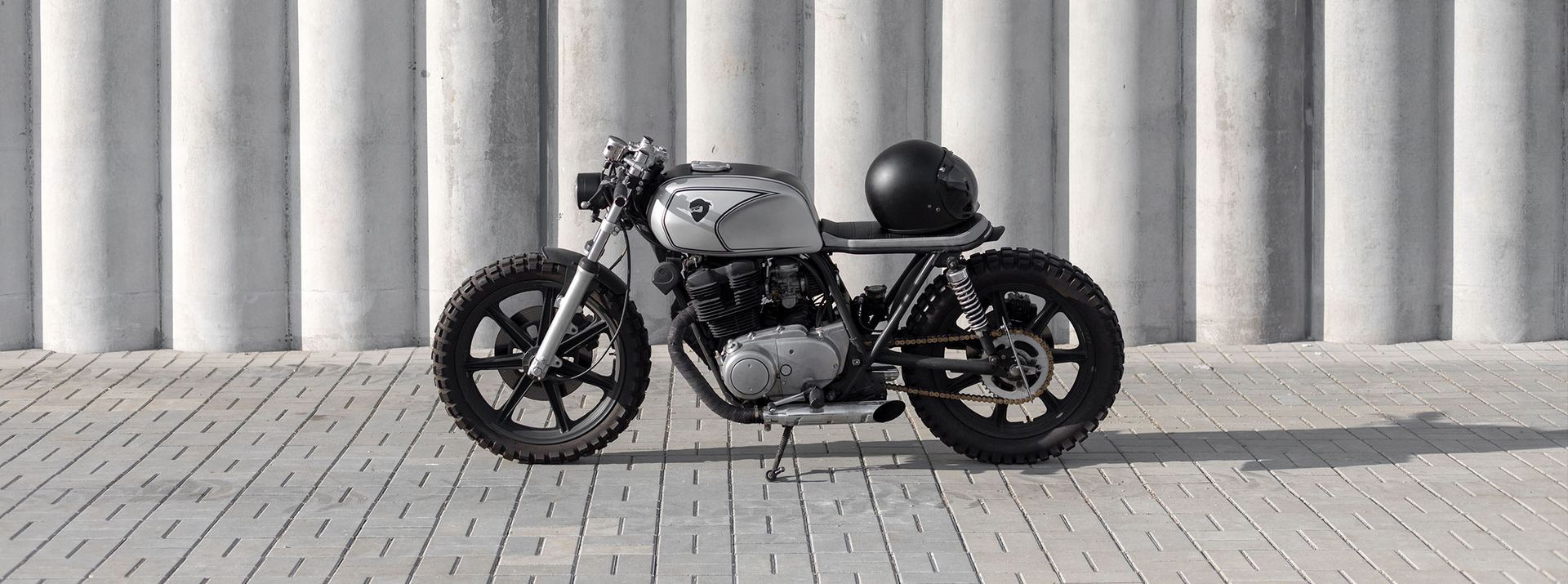 A silver cafe racer motorcycle with a black helmet, parked in front of a concrete wall.