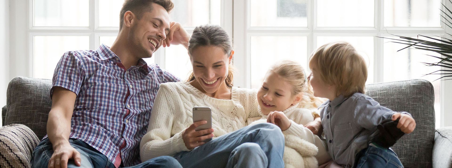 A family of four looking at a smartphone together on a couch, smiling.