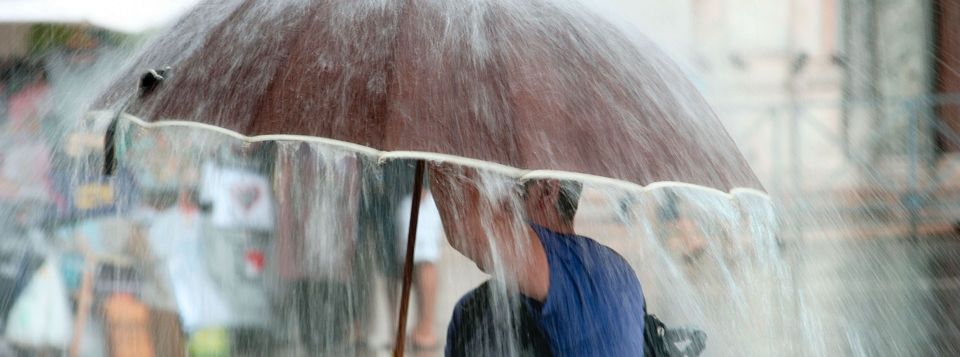 Person under a brown umbrella in the rain. Water pours down, blurring the background.