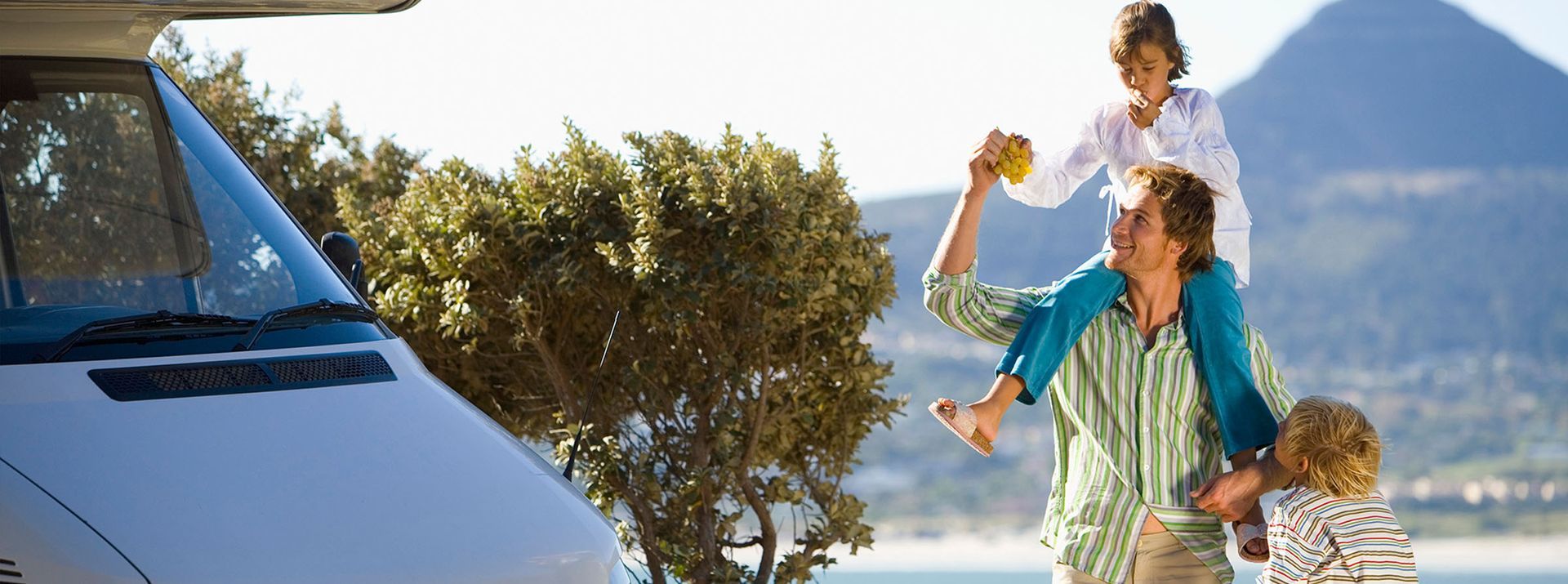 Family enjoying outdoors: man carrying child on shoulders, child holding up food, RV, mountains in background.