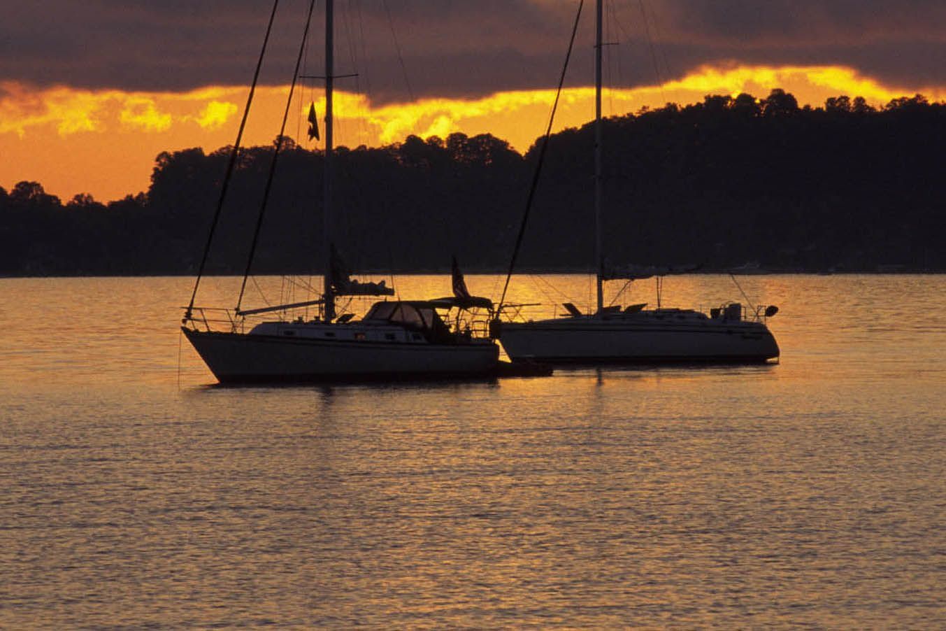 Two sailboats on calm water at sunset, silhouetted against a golden and orange sky.