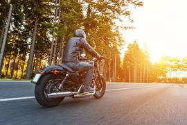Motorcyclist riding on a road through a sunny forest, wearing a black leather jacket.