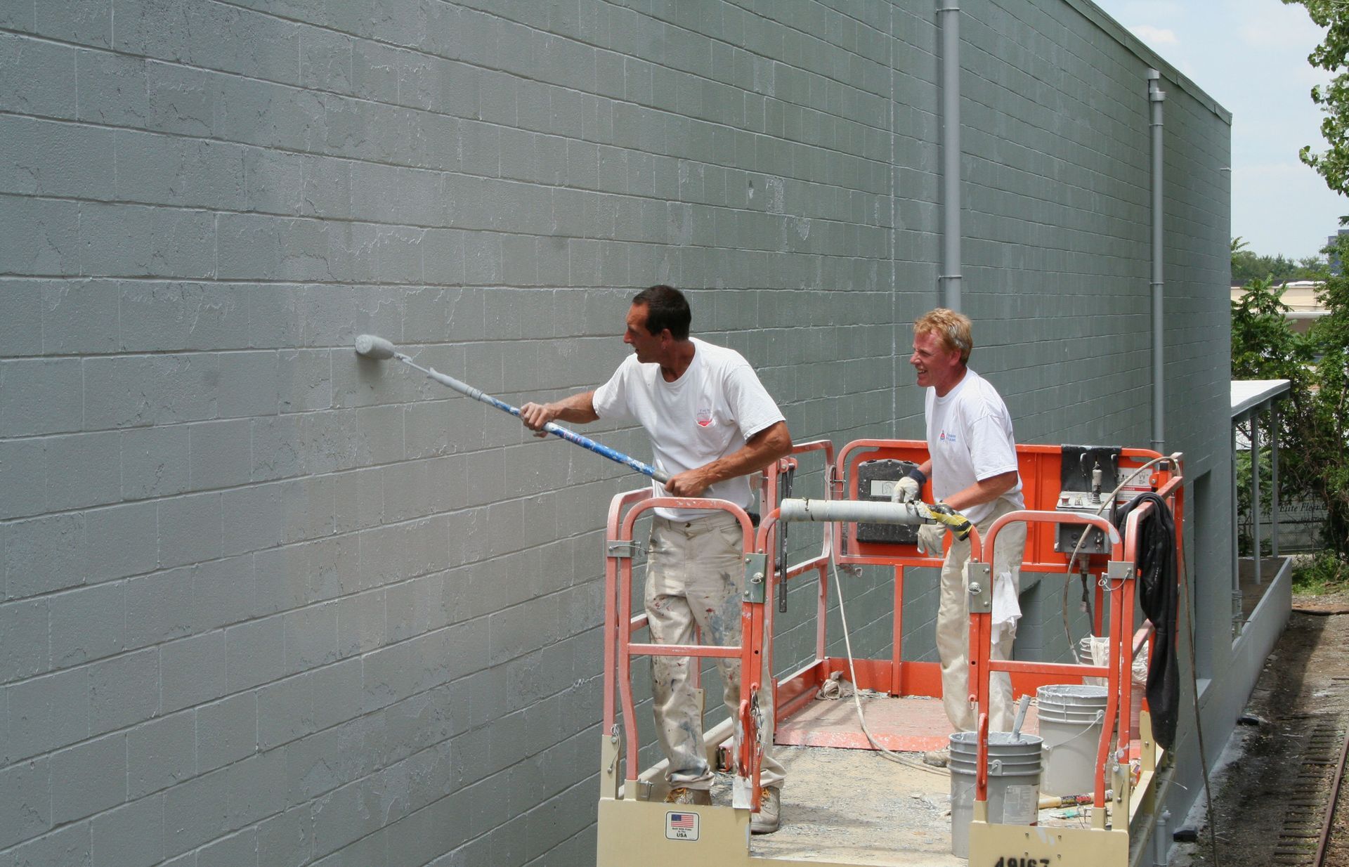 Two workers on a lift painting a gray exterior wall with a spray roller