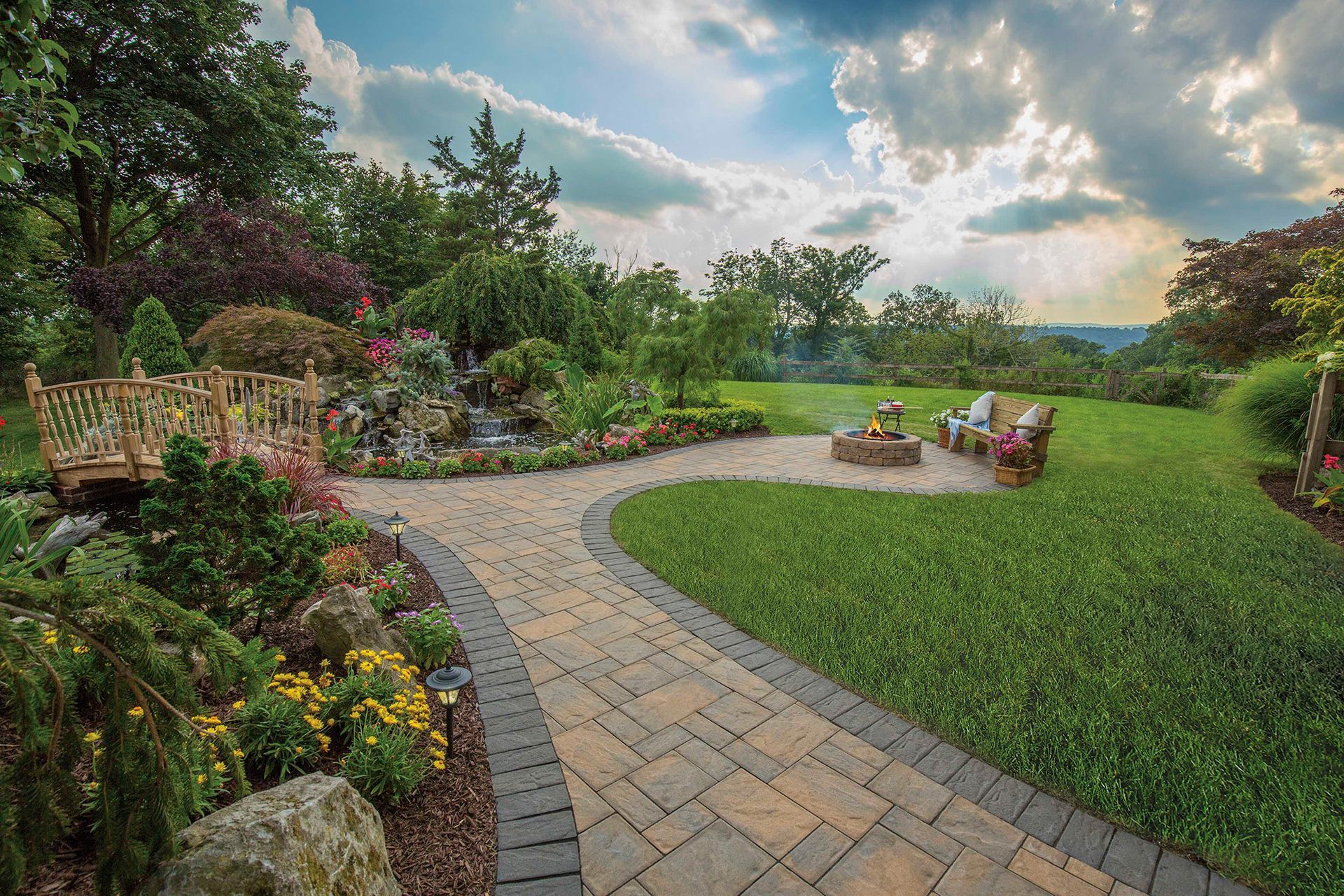 A brick walkway leading to a fire pit in a lush green garden.