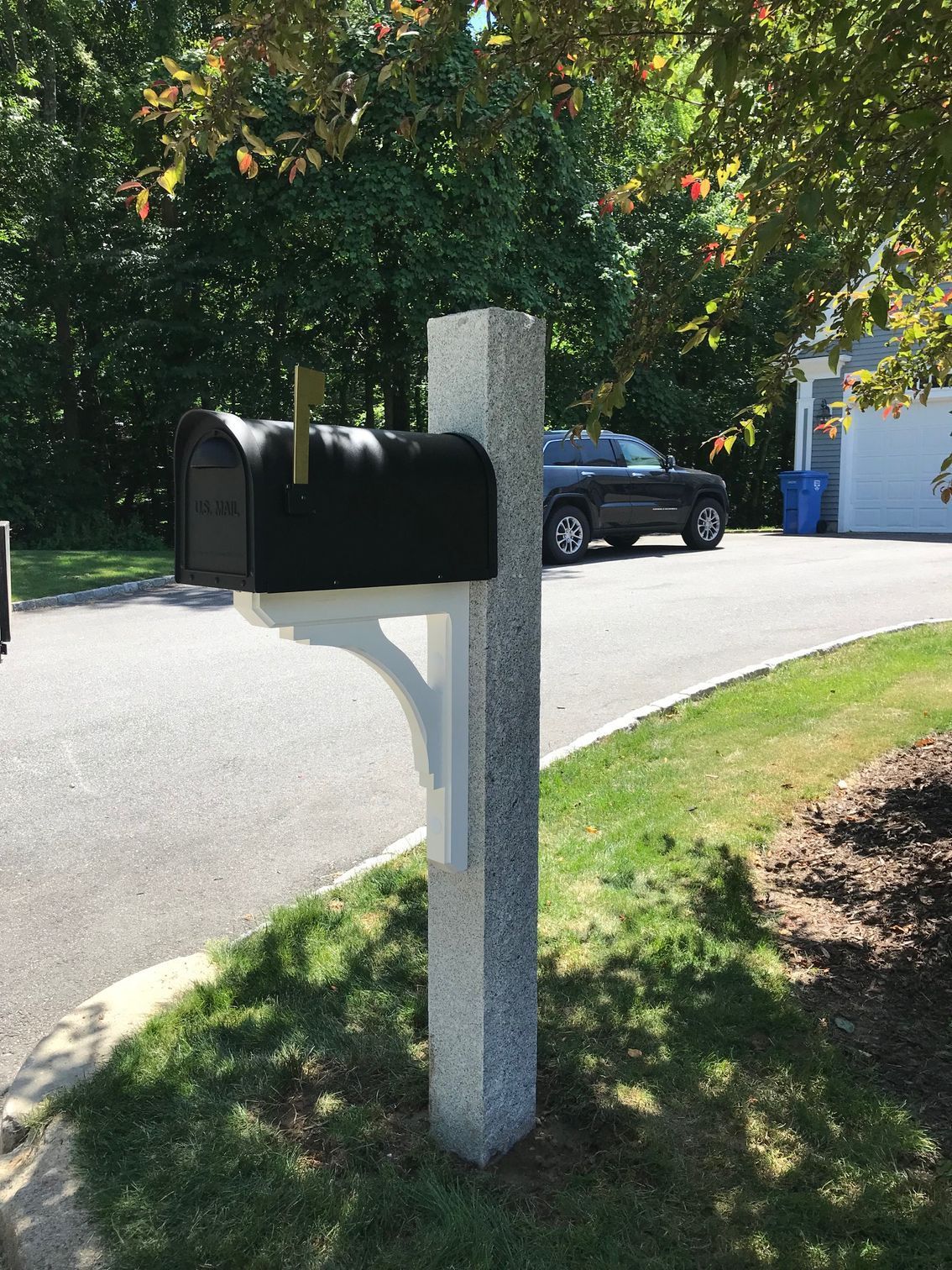 A black mailbox mounted on a white bracket attached to a grey, textured stone post beside a paved driveway.