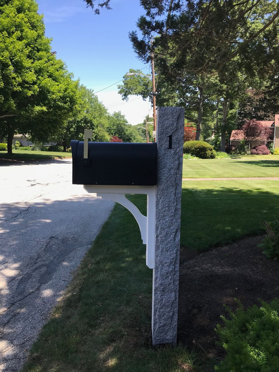A black mailbox attached to a gray stone post with a white wooden support bracket, situated beside a suburban street.
