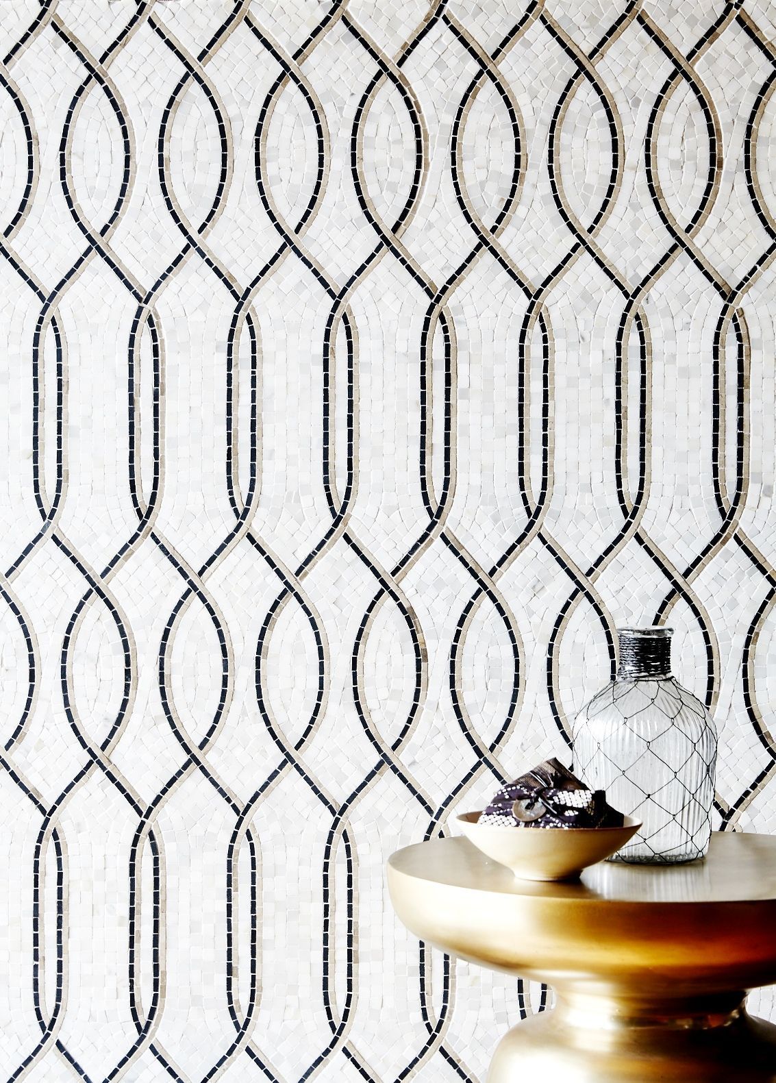 There is a vase on a gold plate in front of a wall with a pattern on it