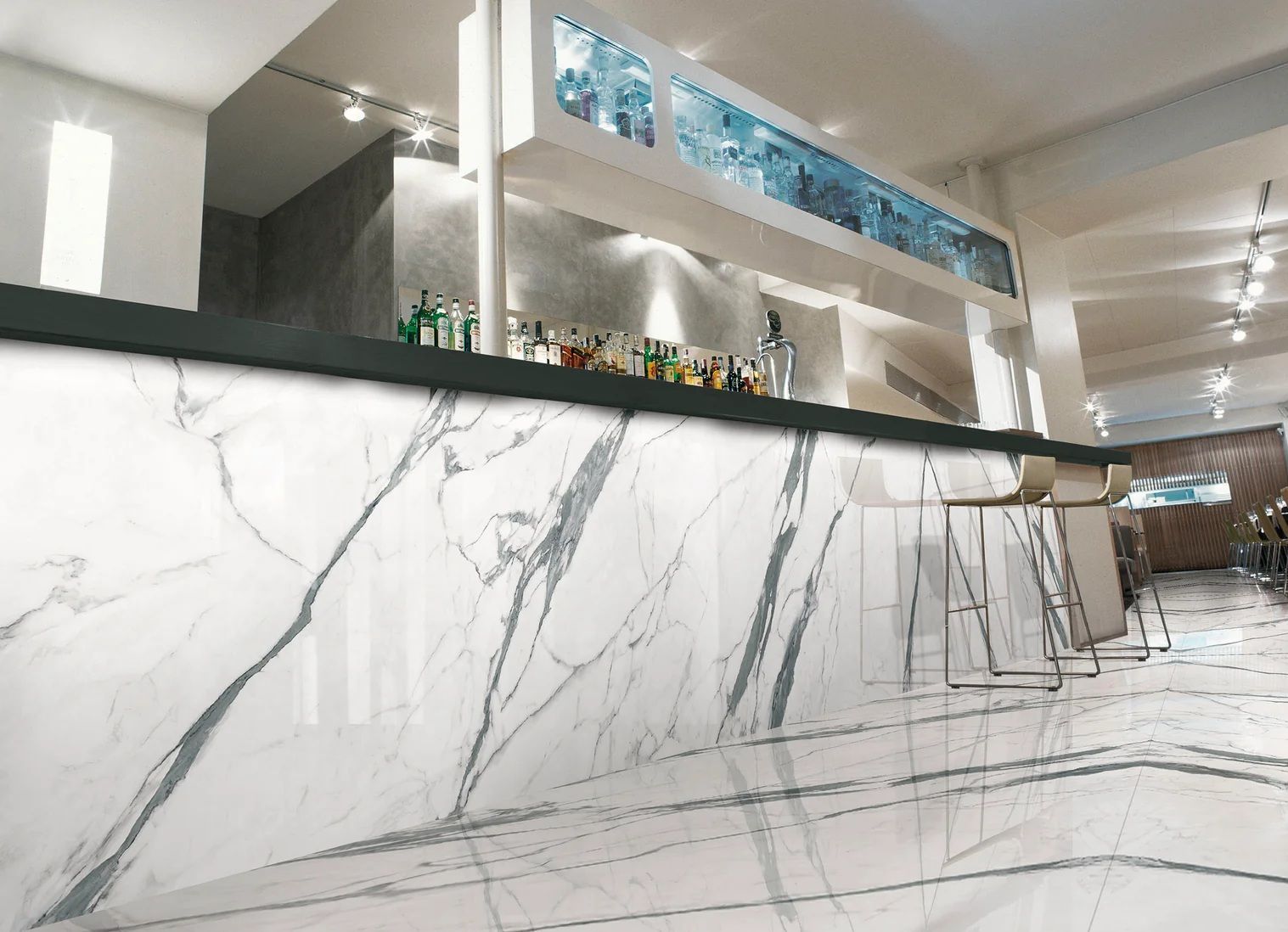 Bar with white marble facade and counter, bottles and bar stools. Brightly lit interior.