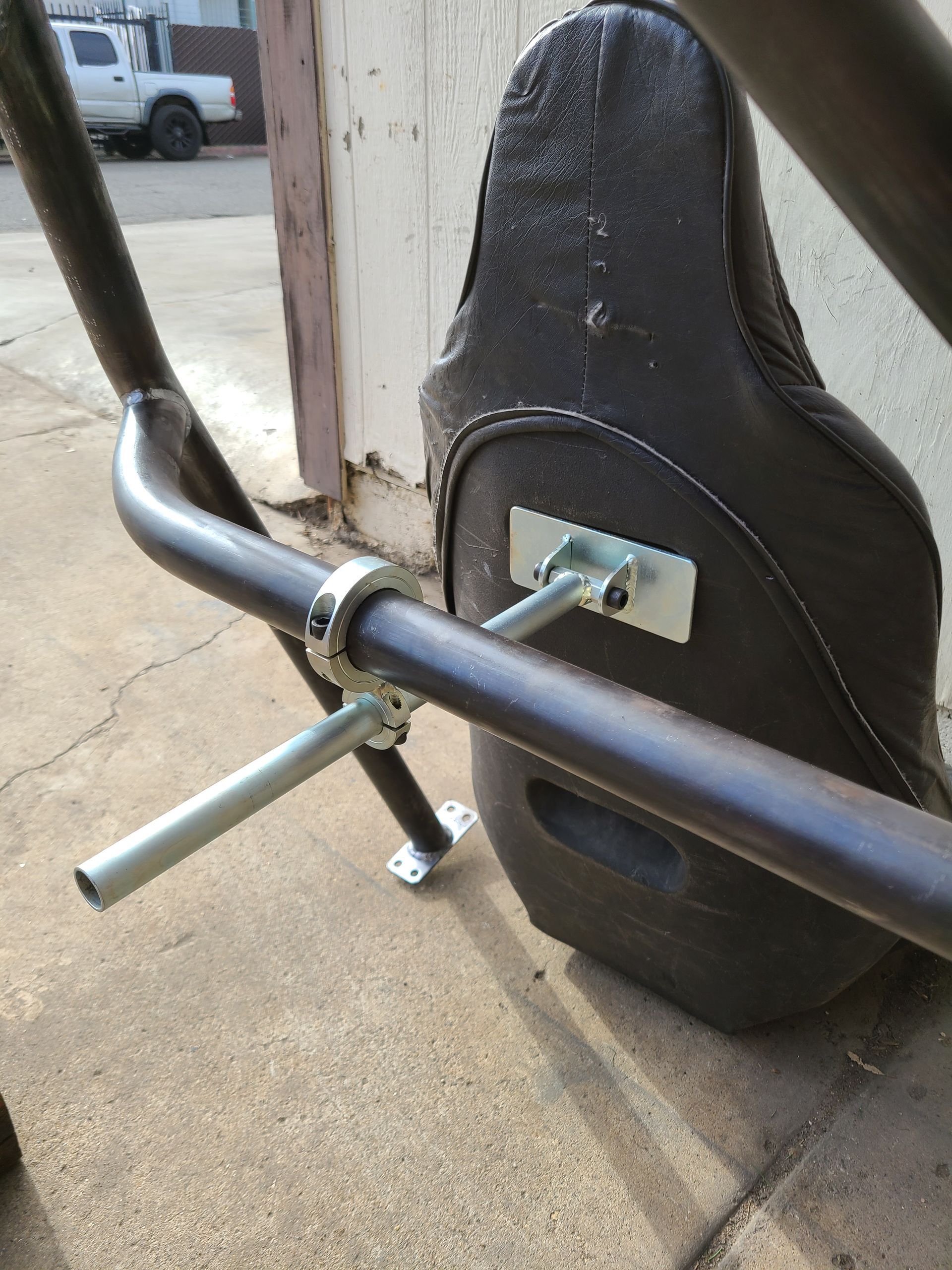 Brown racing seat attached to a roll cage with metal brackets and clamps on a concrete surface.