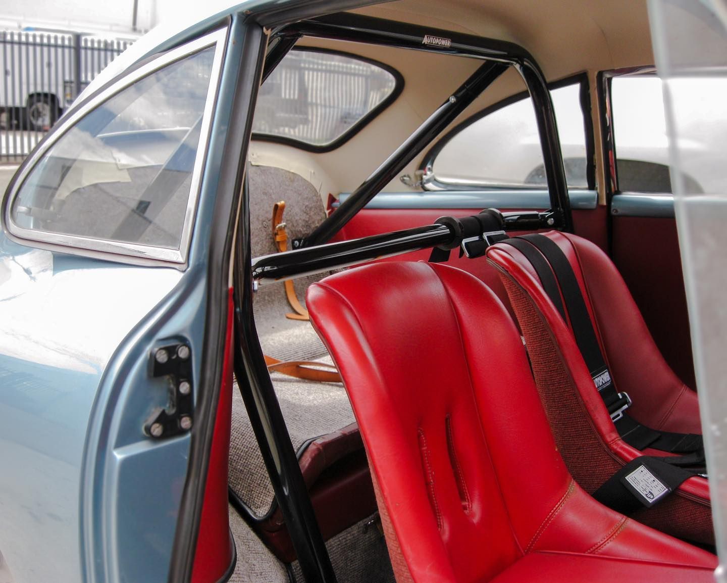 Interior of a vintage light blue car with red racing seats and a black roll cage.