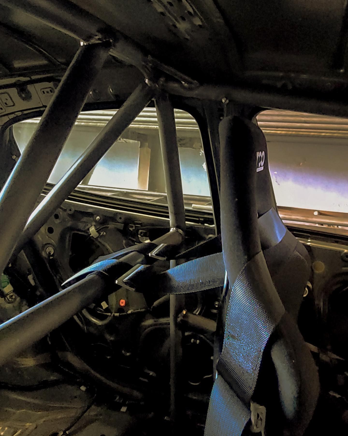 Inside of a race car: roll cage, racing seats, black belts, and exposed metal frame.