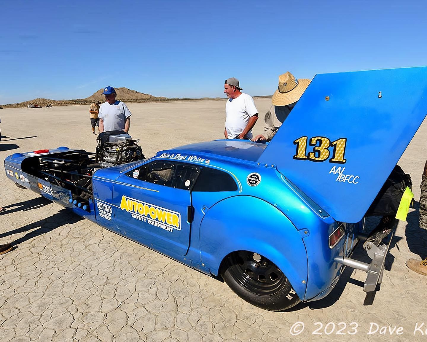 Blue race car, number 131, on a dry lake bed; crew members nearby with open hood.