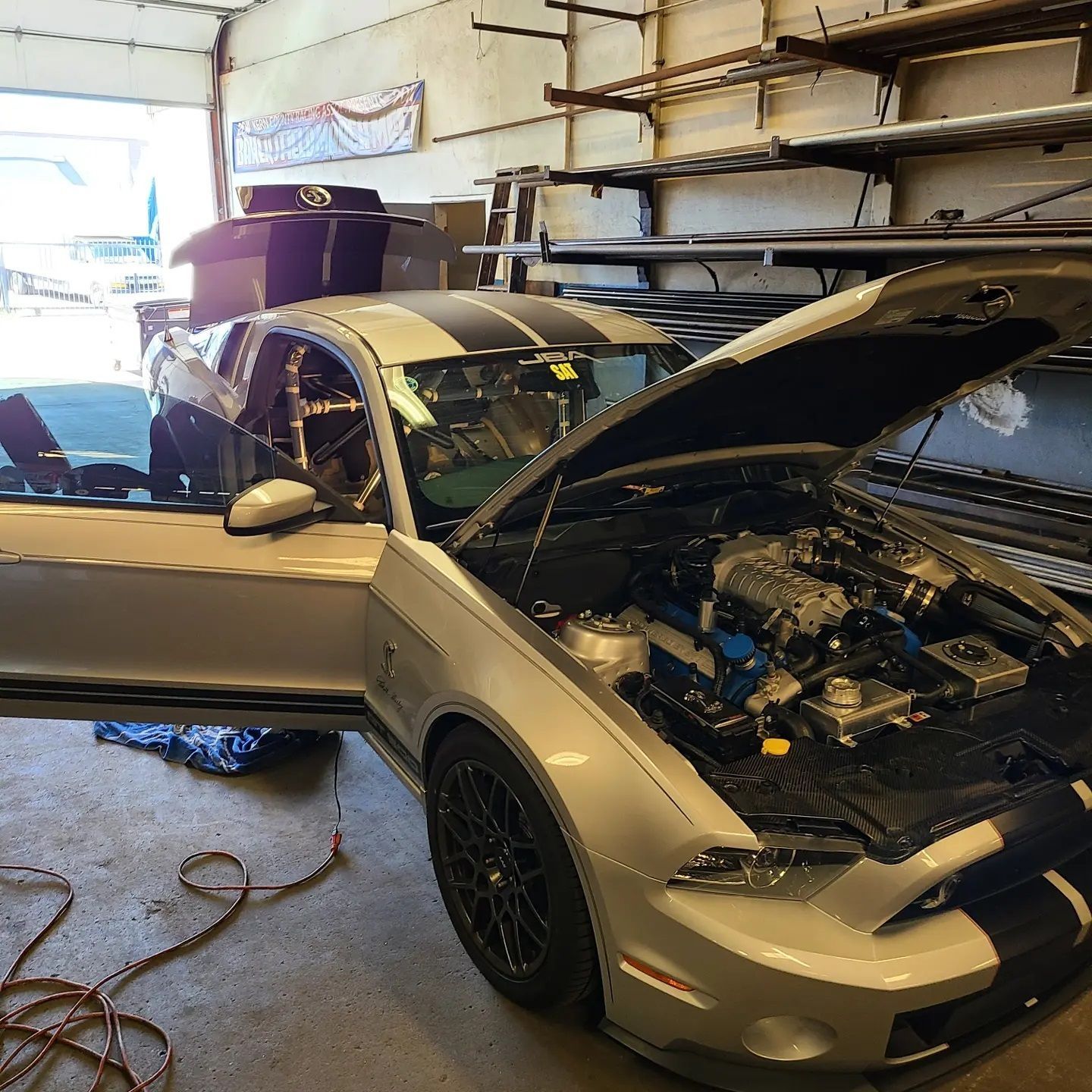 Silver Mustang with black racing stripes and open hood in a garage.