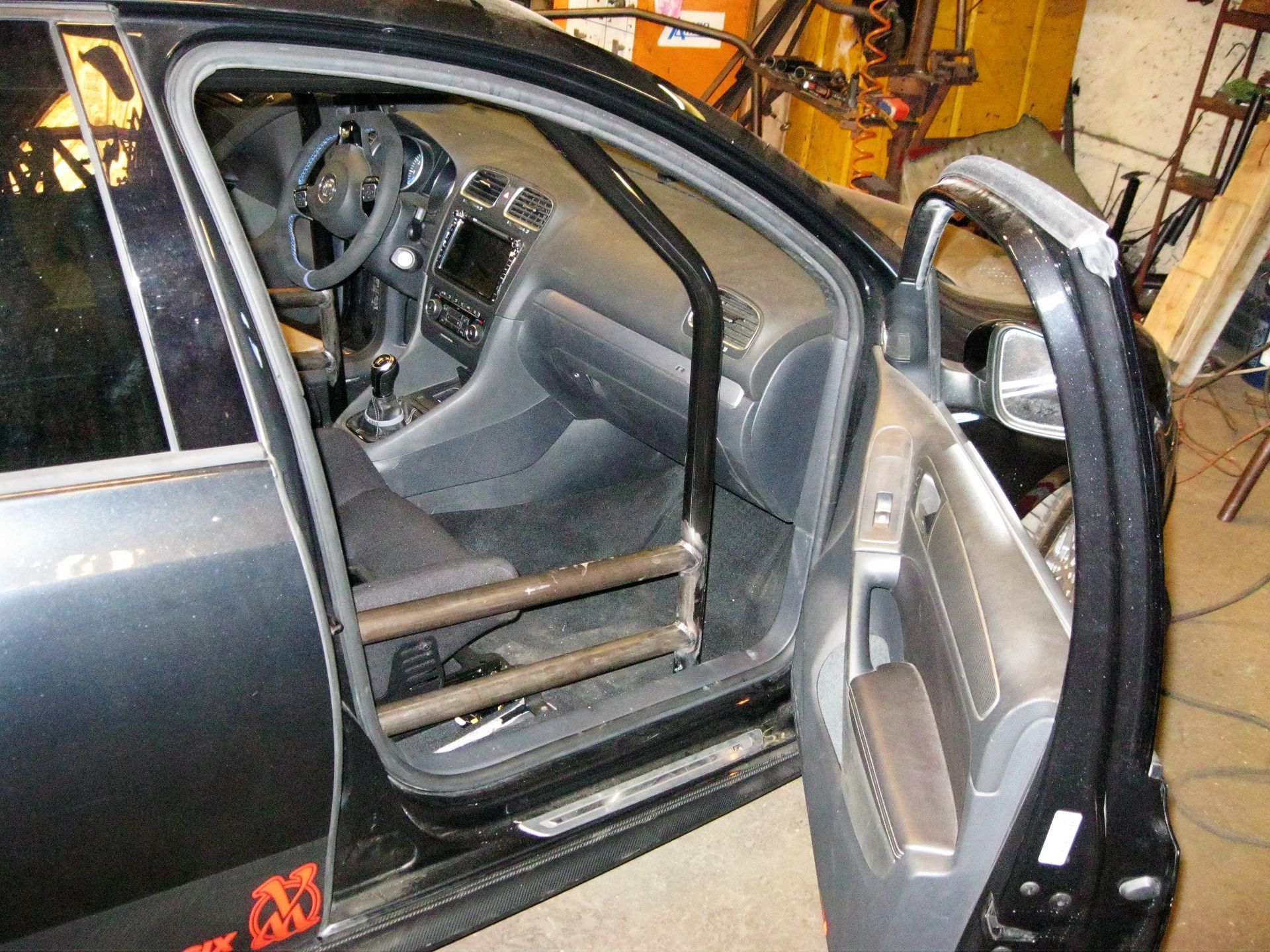 Black car interior with roll cage. Door open, showing dashboard, steering wheel, and racing seat.