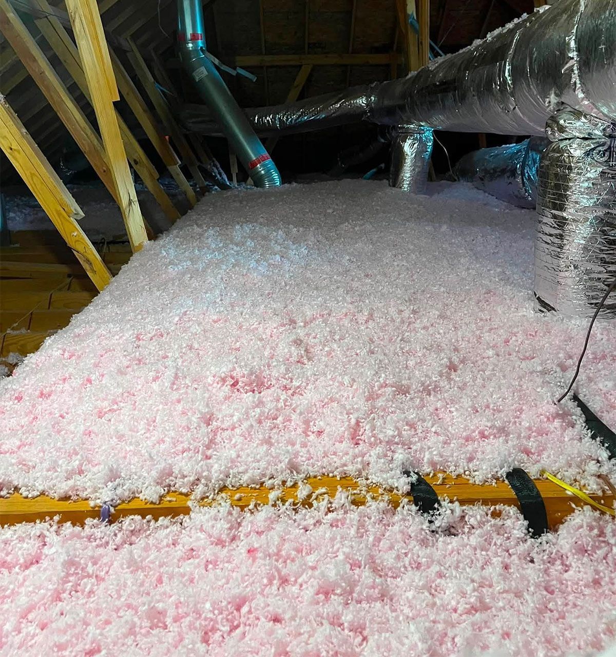 Attic crawlspace floor covered with pink and white foam beads beside metal ducts and wooden framing