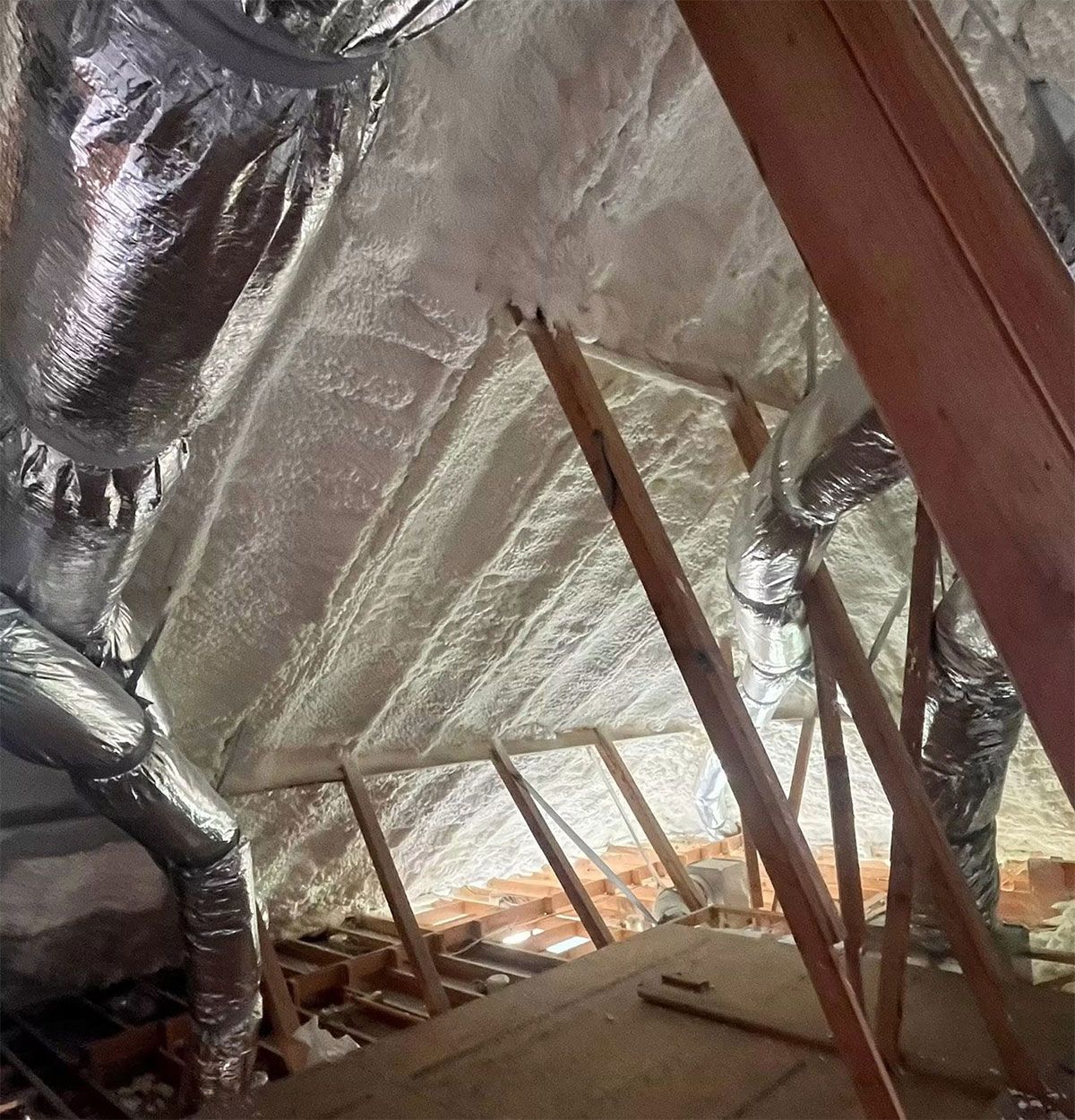 Inside an attic, shiny foil insulation and wooden beams around a narrow floor space.