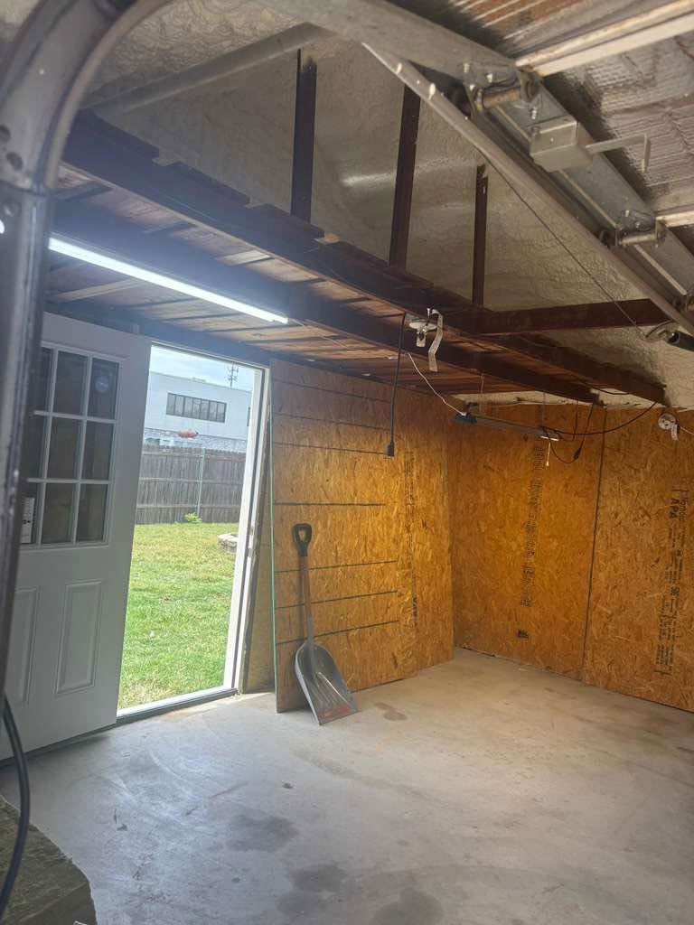 Empty garage interior with open side door, concrete floor, exposed ceiling beams, and wood-paneled wall