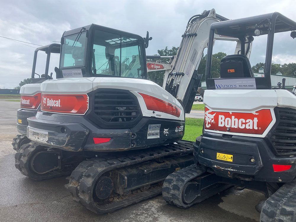Two Bobcat compact excavators parked side by side on wet pavement, red and white one in front.