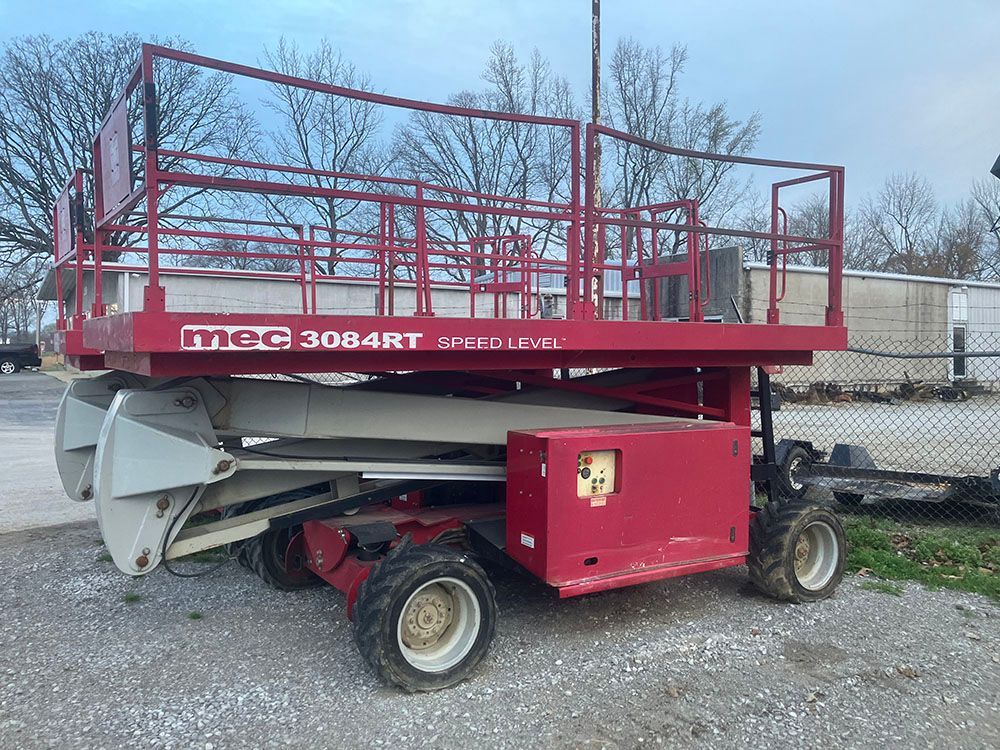 Red MEC 3084RT scissor lift parked outdoors on gravel near a fence.