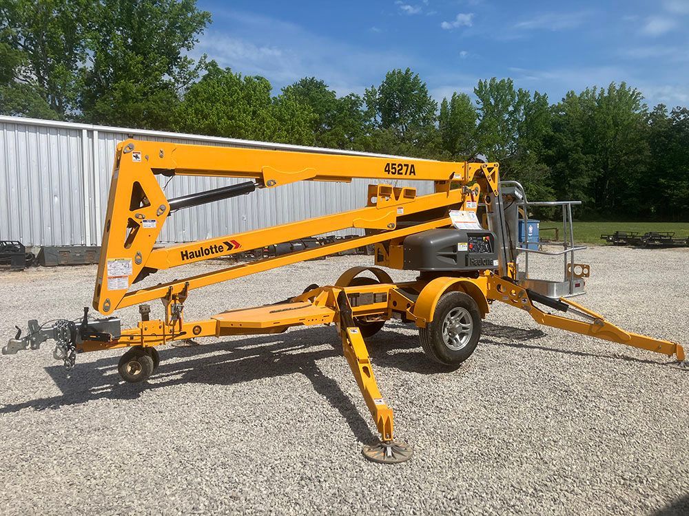 Yellow towable boom lift on gravel lot, with outriggers extended beside a white building and trees.