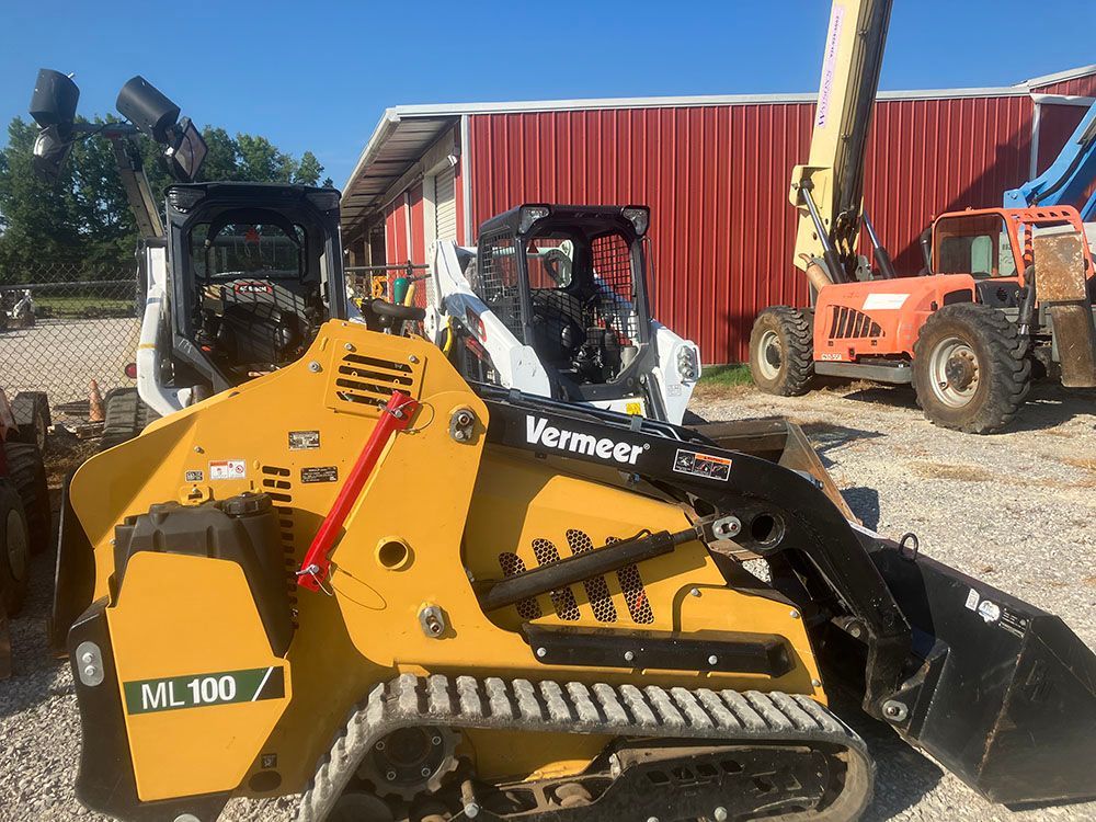 Yellow Vermeer mini skid steer with bucket in a lot beside red barn and other construction equipment.