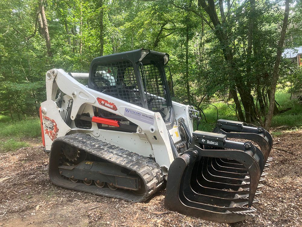 White compact track loader with grapple bucket parked on a dirt path in a wooded area.