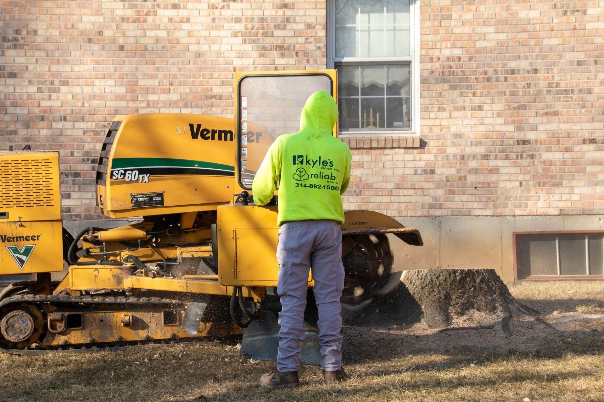 Person operating a yellow stump grinder next to a brick building.