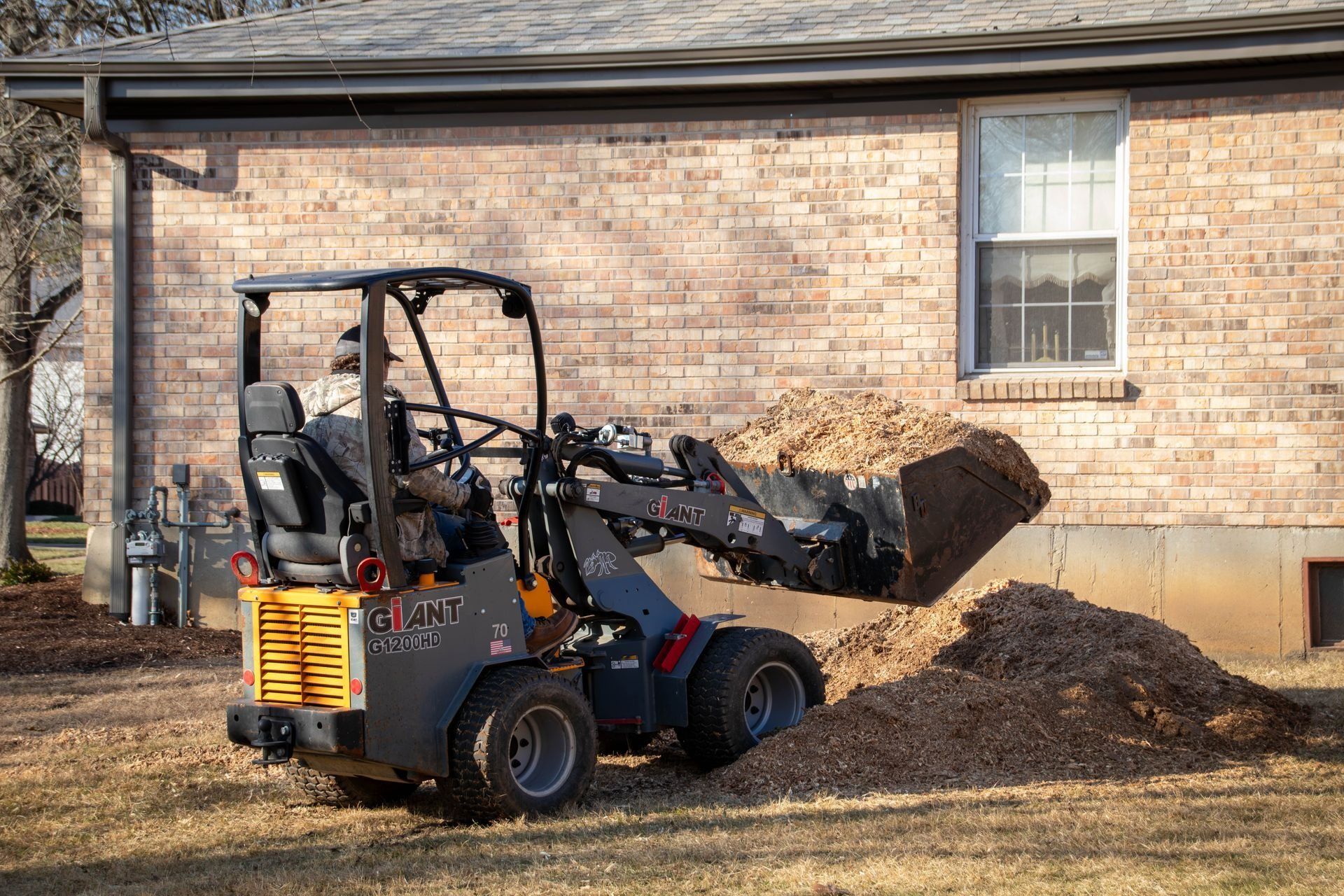 Small yellow and grey loader moving mulch near a brick building.