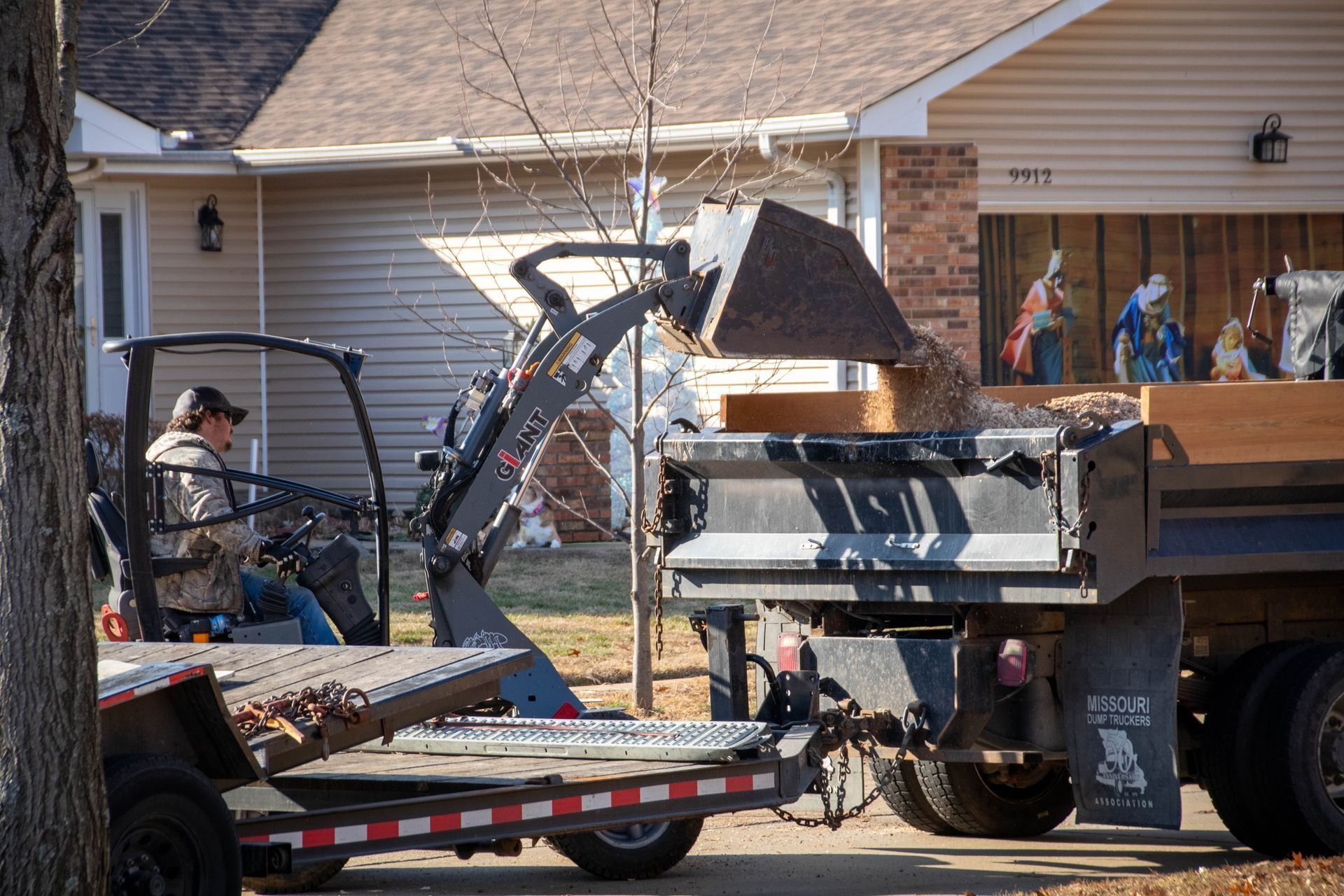A person in a tractor loads wood into a truck in front of a house.