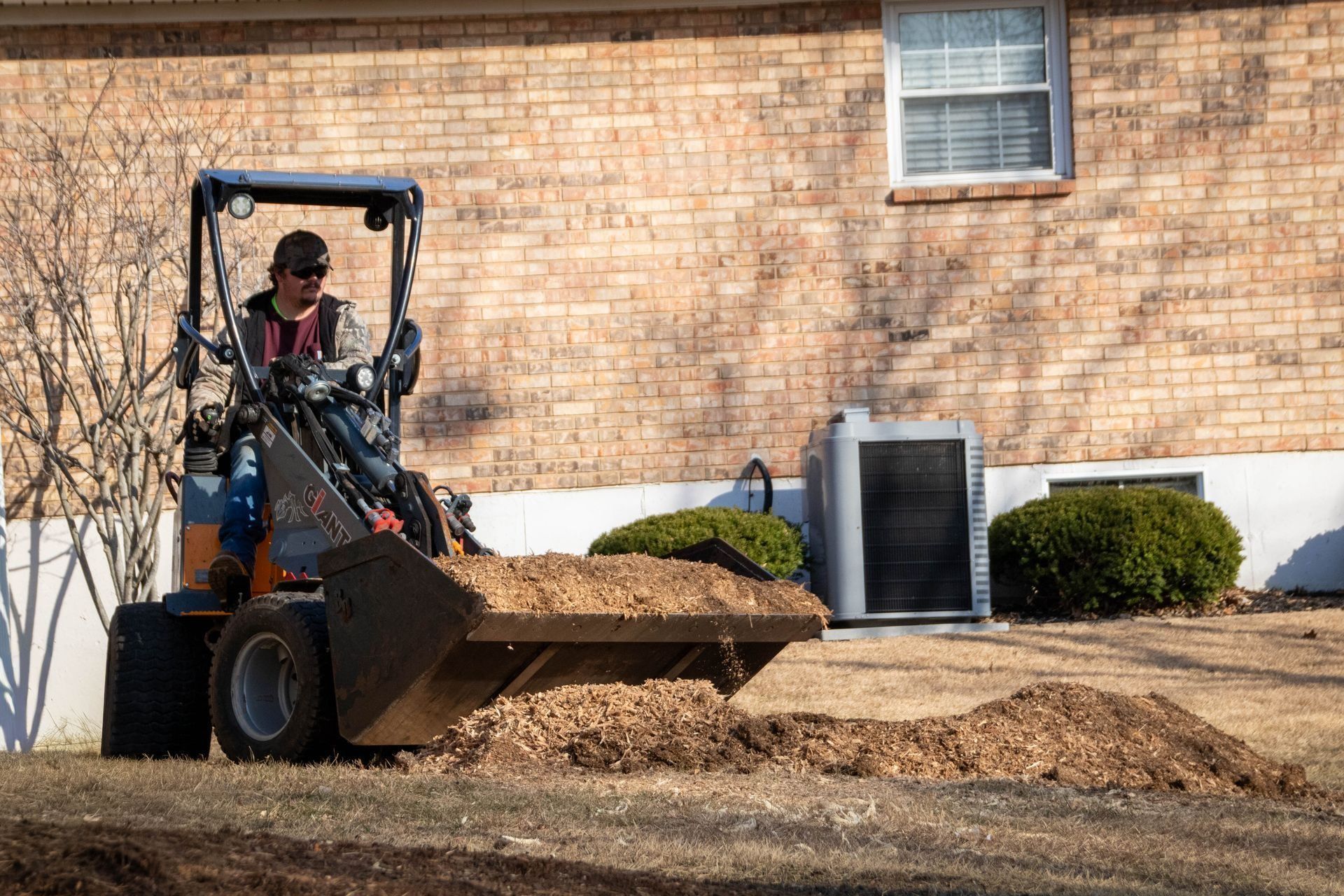 A person operates a small front loader, moving a pile of mulch in front of a brick building.