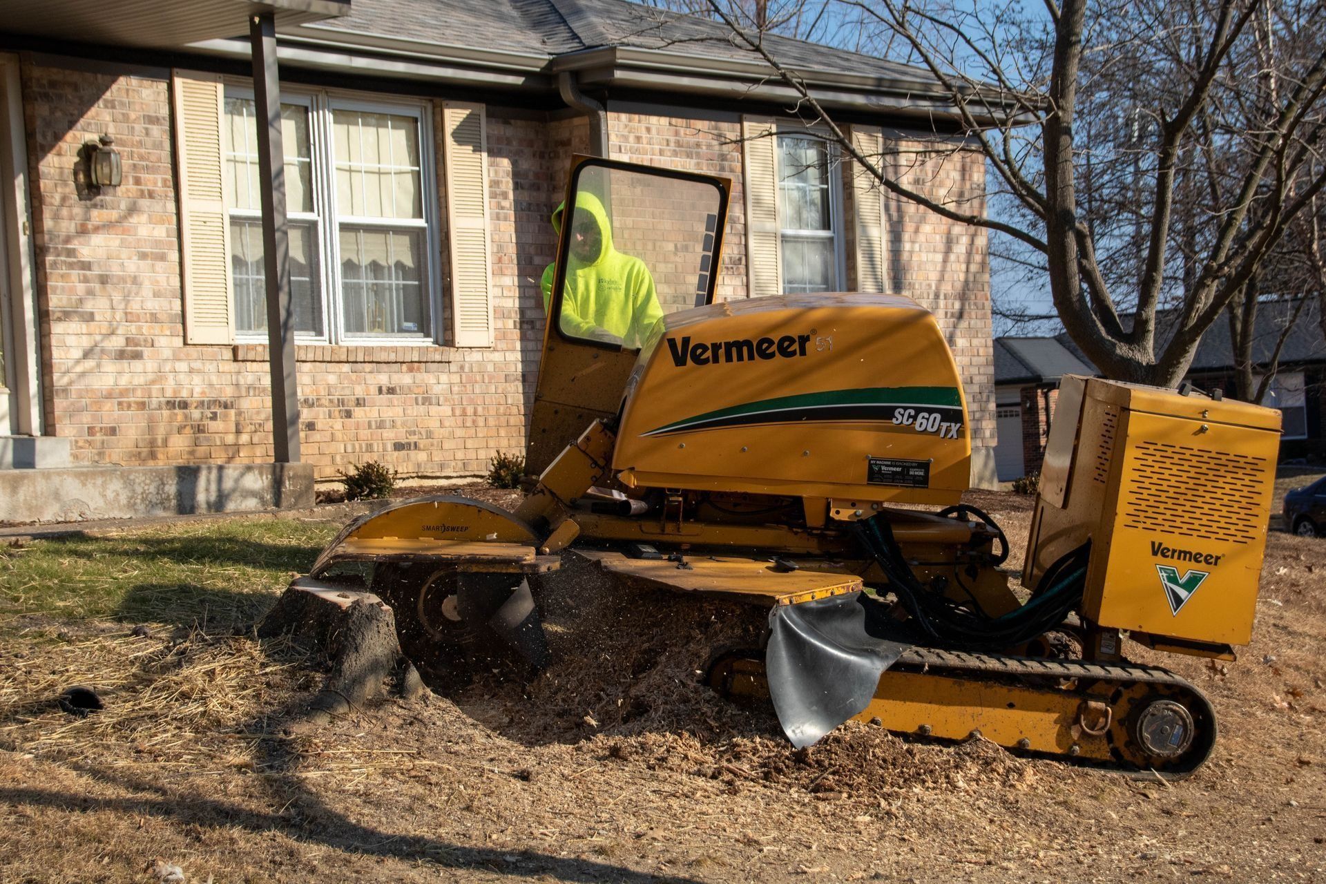 Yellow stump grinder removing a tree stump in front of a brick house. Person in safety vest operates machine.