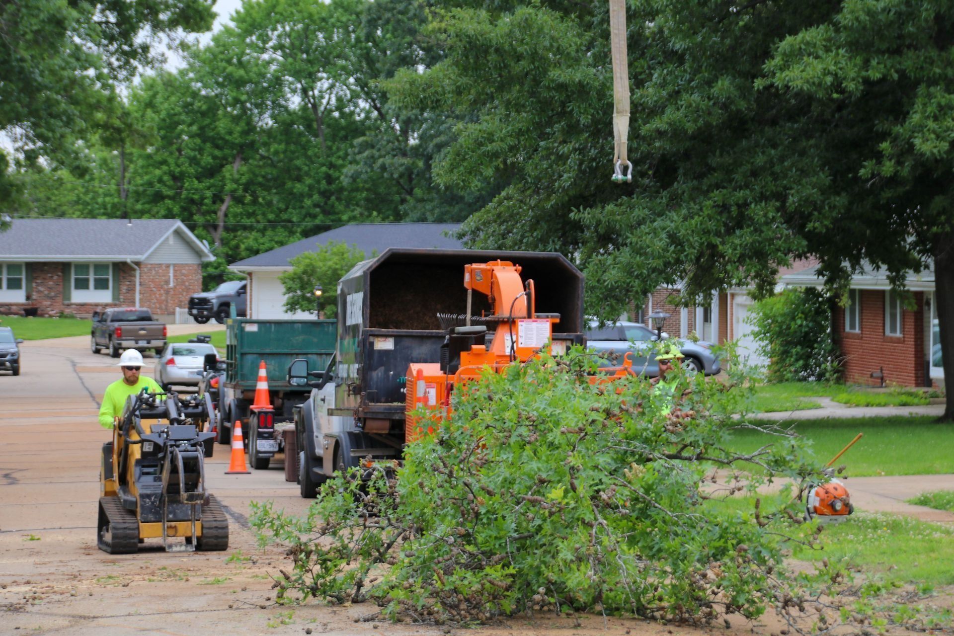 Workers clearing tree debris with chipper truck and mini excavator on a residential street.