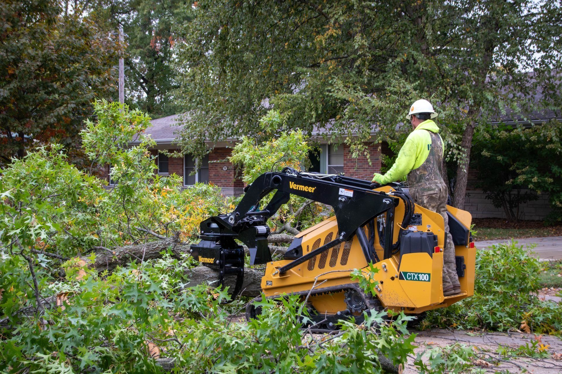 Man operating a yellow skid-steer with a tree shear, cutting branches. Outdoors in front of a house.