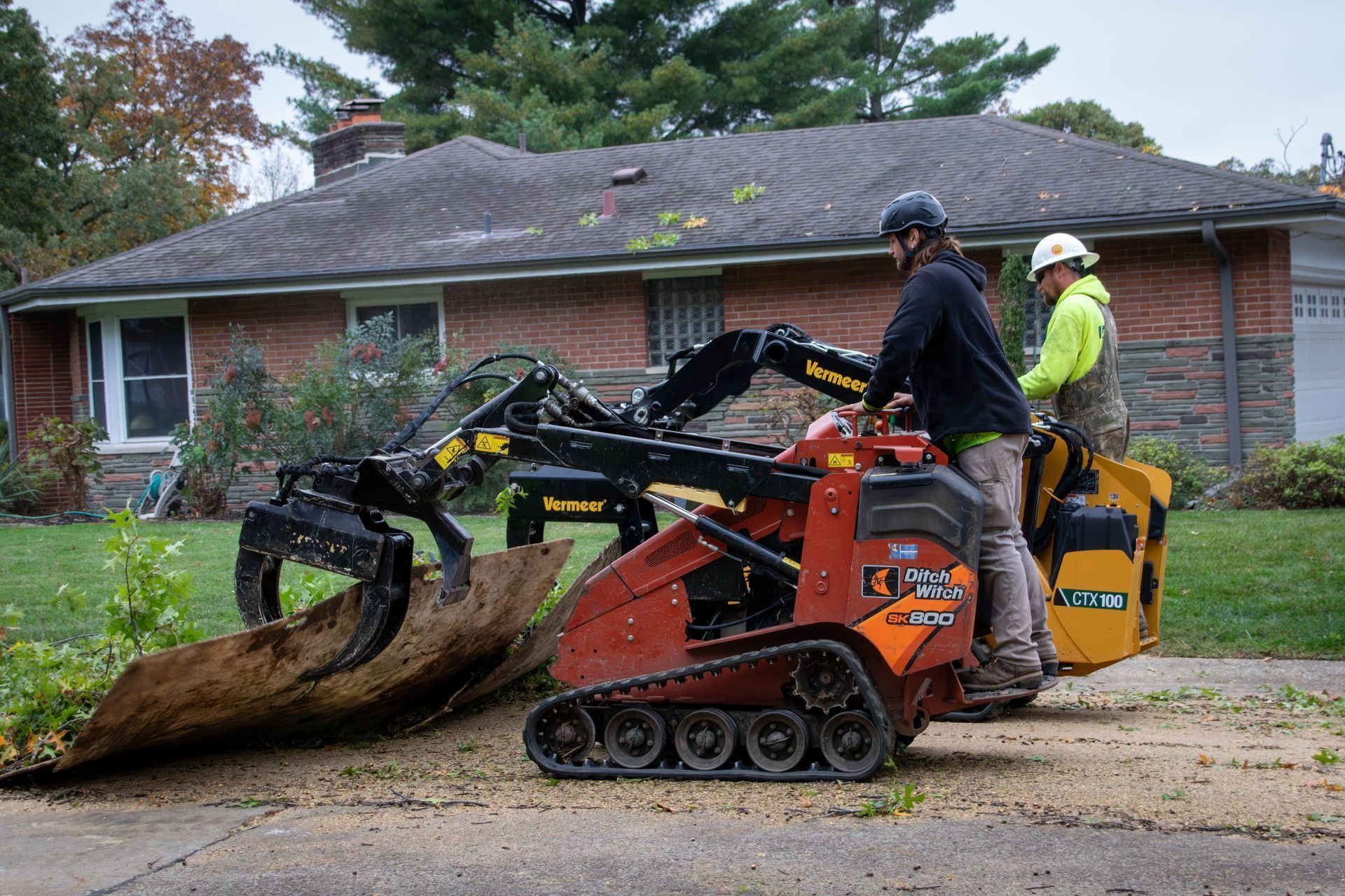 Two workers operating a mini excavator to remove debris in front of a house.