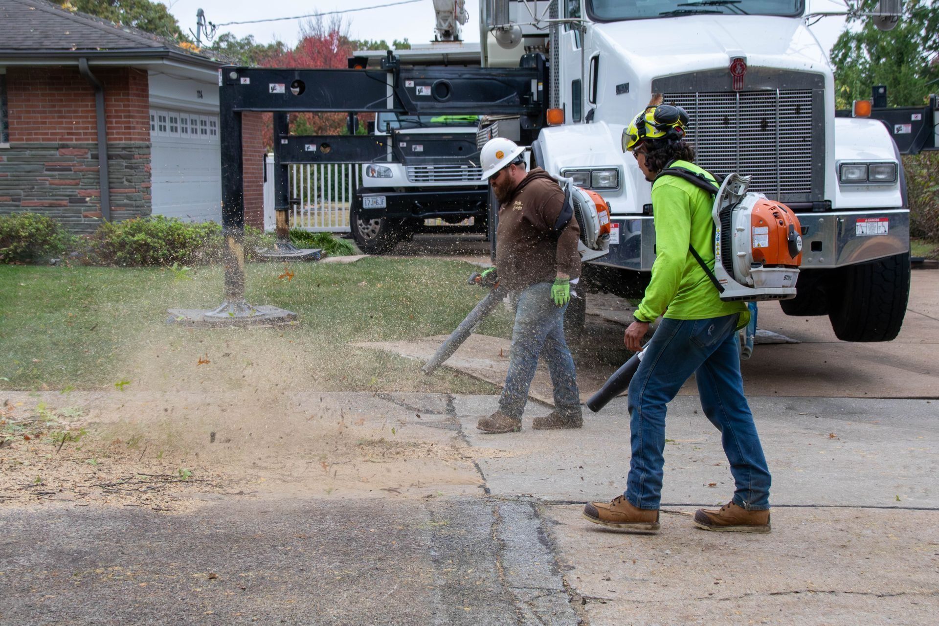 Two workers using leaf blowers to clear debris from a sidewalk near a truck, brick house in background.