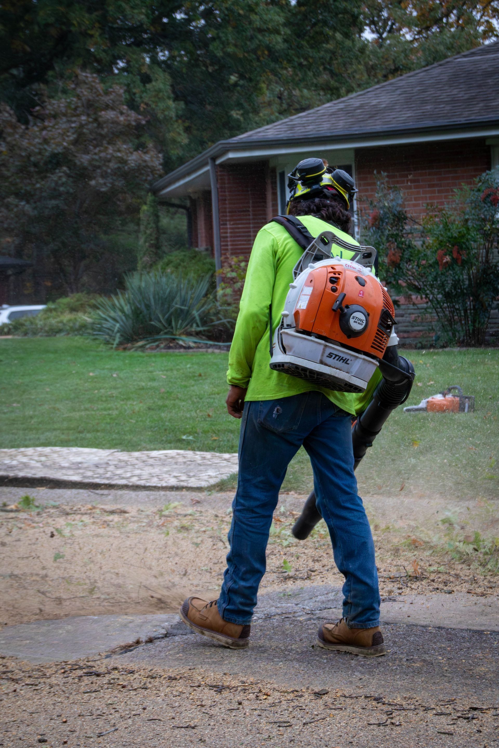 Person in neon green shirt and jeans using a backpack leaf blower on a driveway in front of a house.