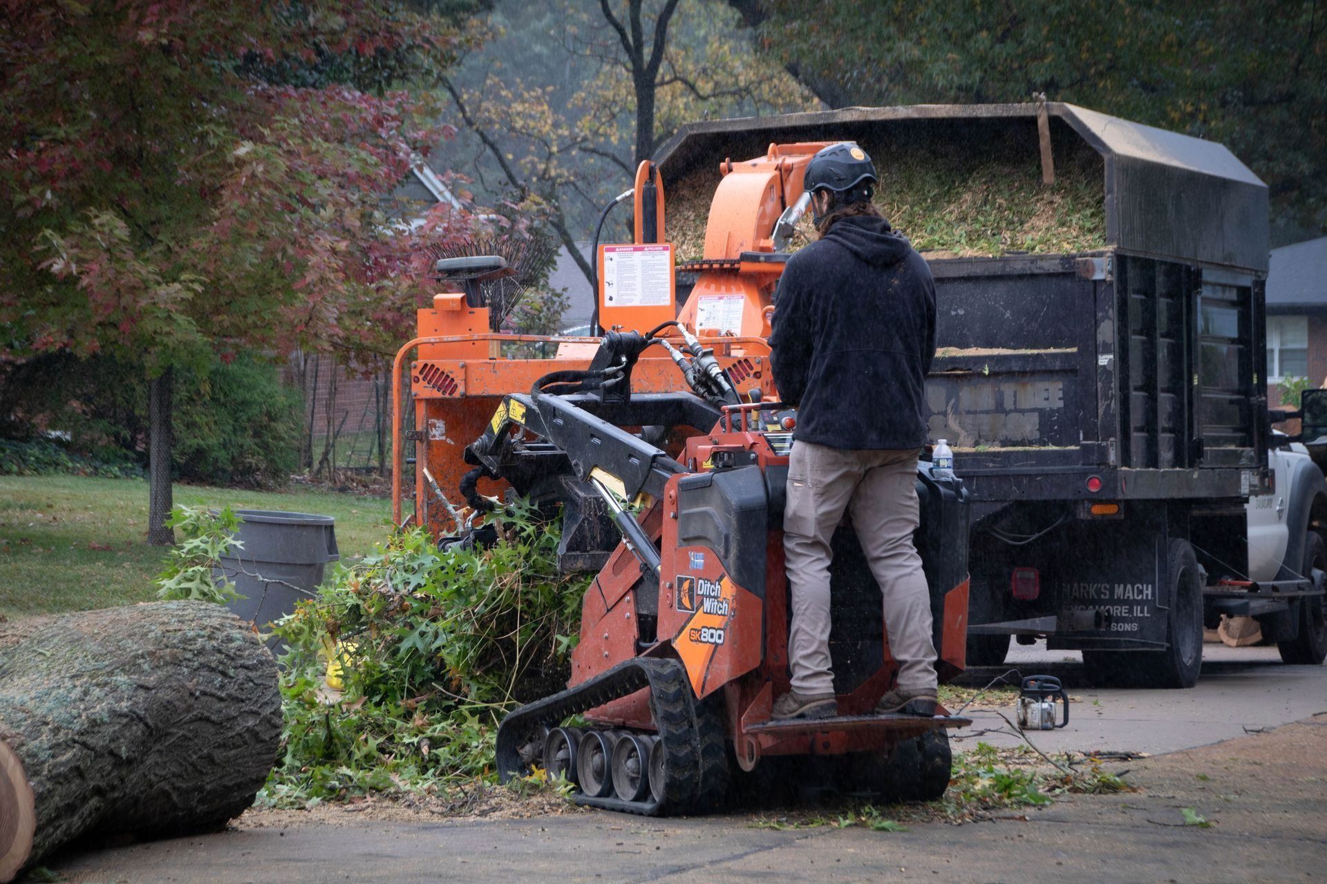 Man operating wood chipper, feeding branches. Orange machine, truck in background, residential setting.