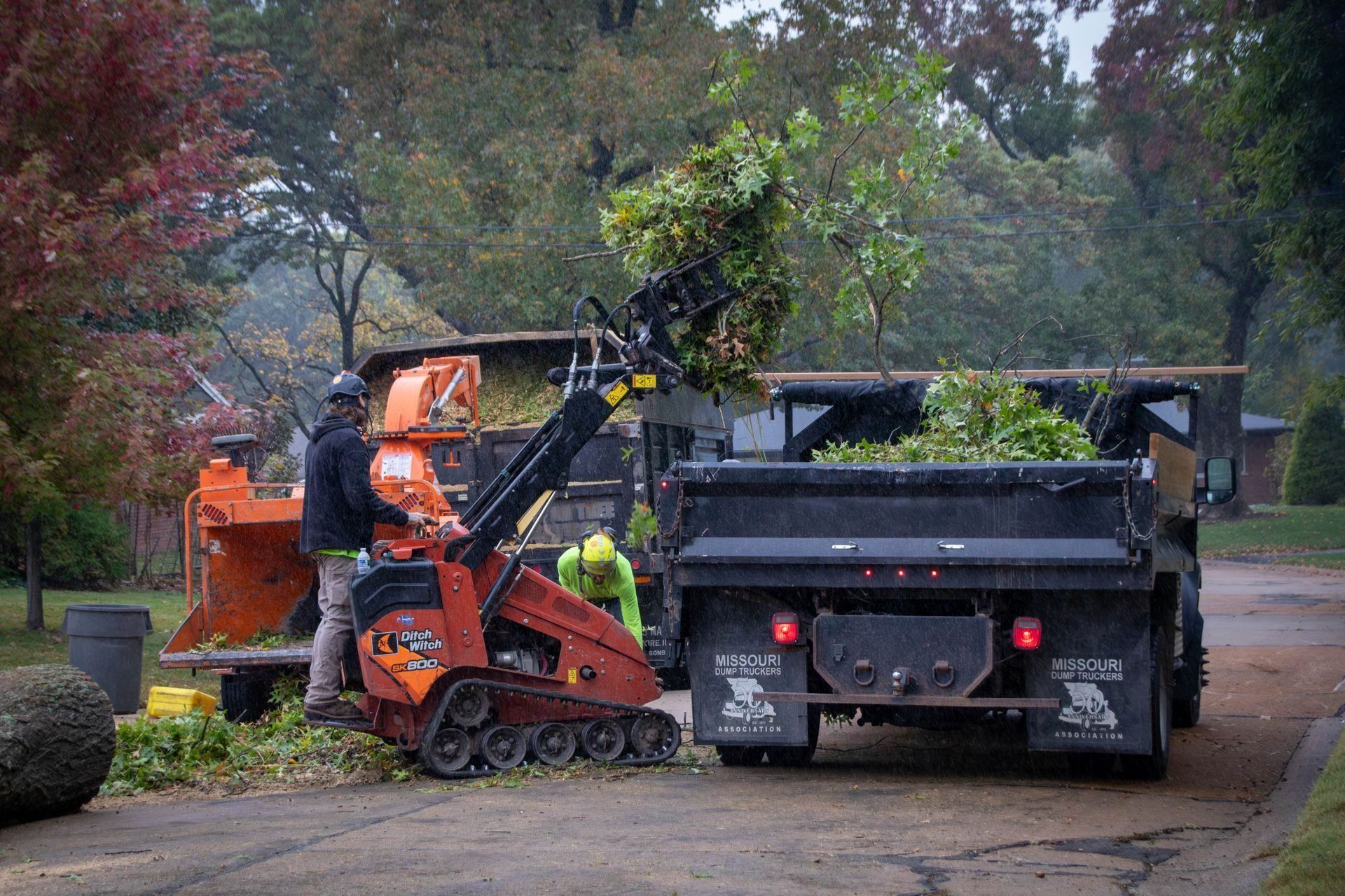 Workers loading tree trimmings into a truck with a chipper on a road.
