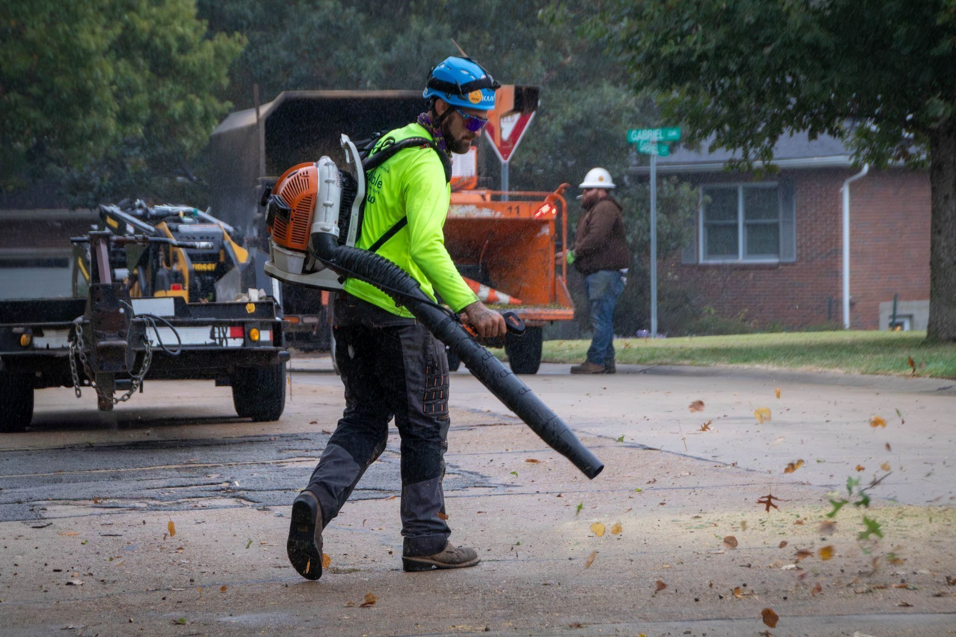 Arborist using a leaf blower on a street, blowing leaves; a truck, chipper, and another worker are visible in the background.
