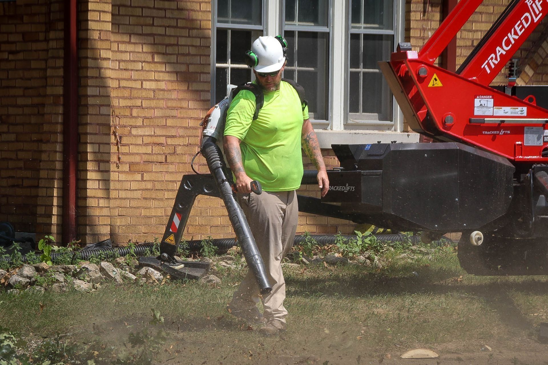 Person using a leaf blower on grass near a red machine and a brick building.