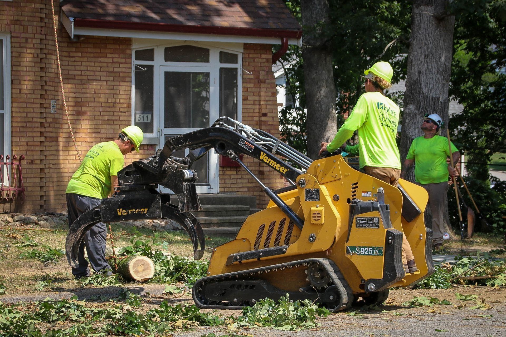 Three workers in neon green shirts and hard hats use a yellow skid steer to remove a tree trunk near a brick house.