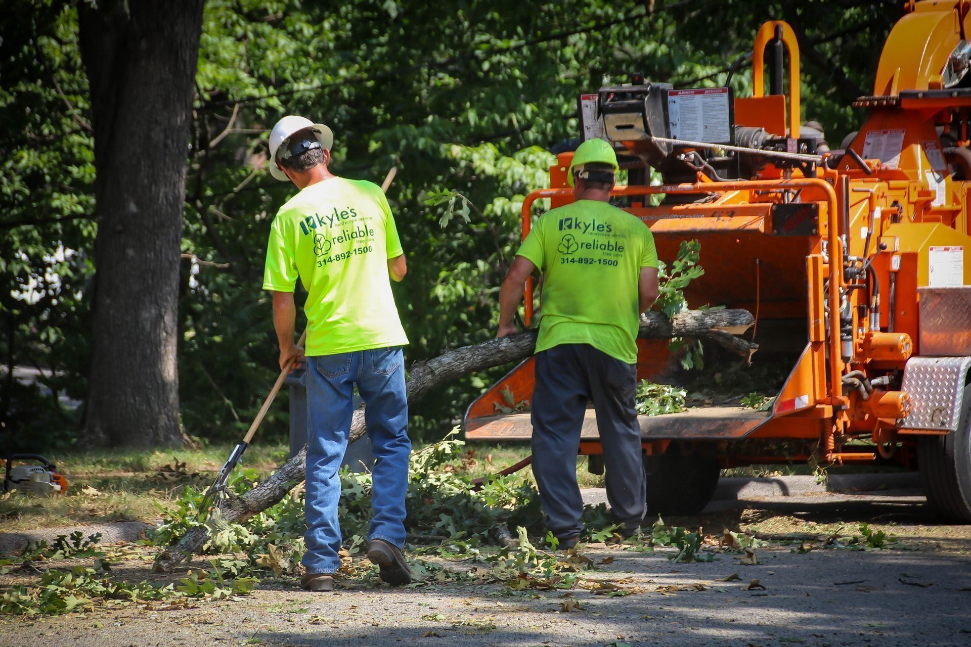Two workers in lime green shirts feeding tree branches into a wood chipper.