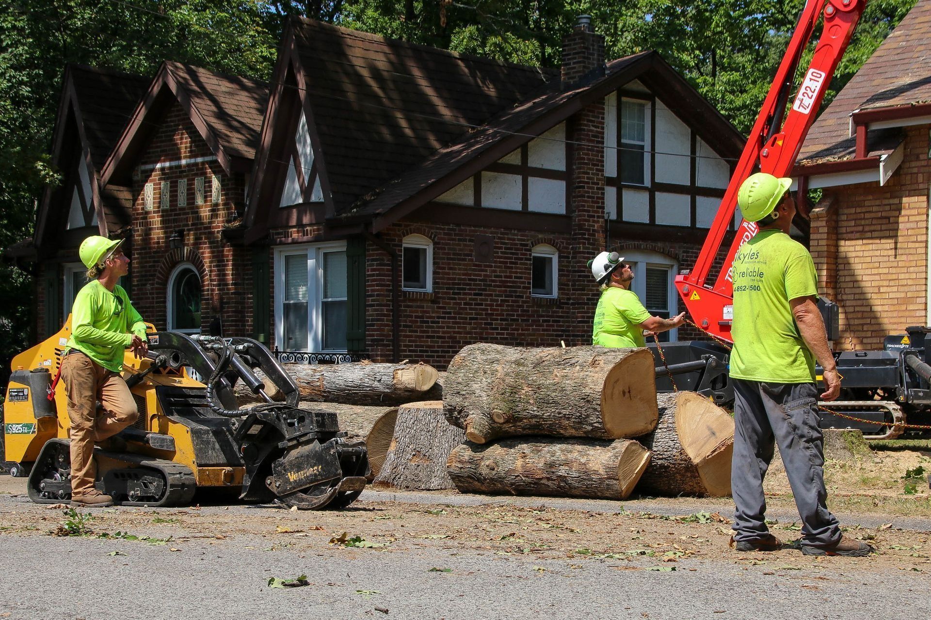 Three tree service workers removing logs near a Tudor-style house with a small skid steer and a crane.
