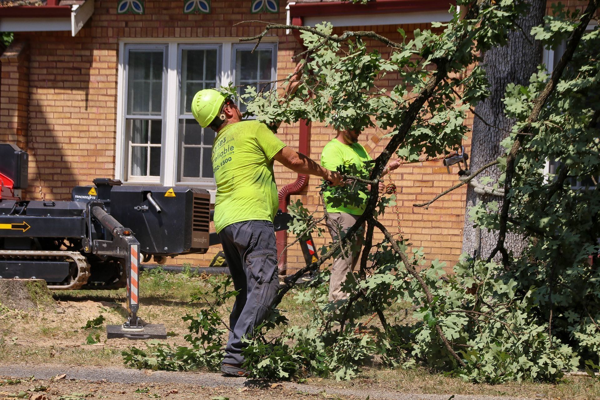 Two workers in neon green shirts trimming a tree in front of a brick house; a wood chipper is present.