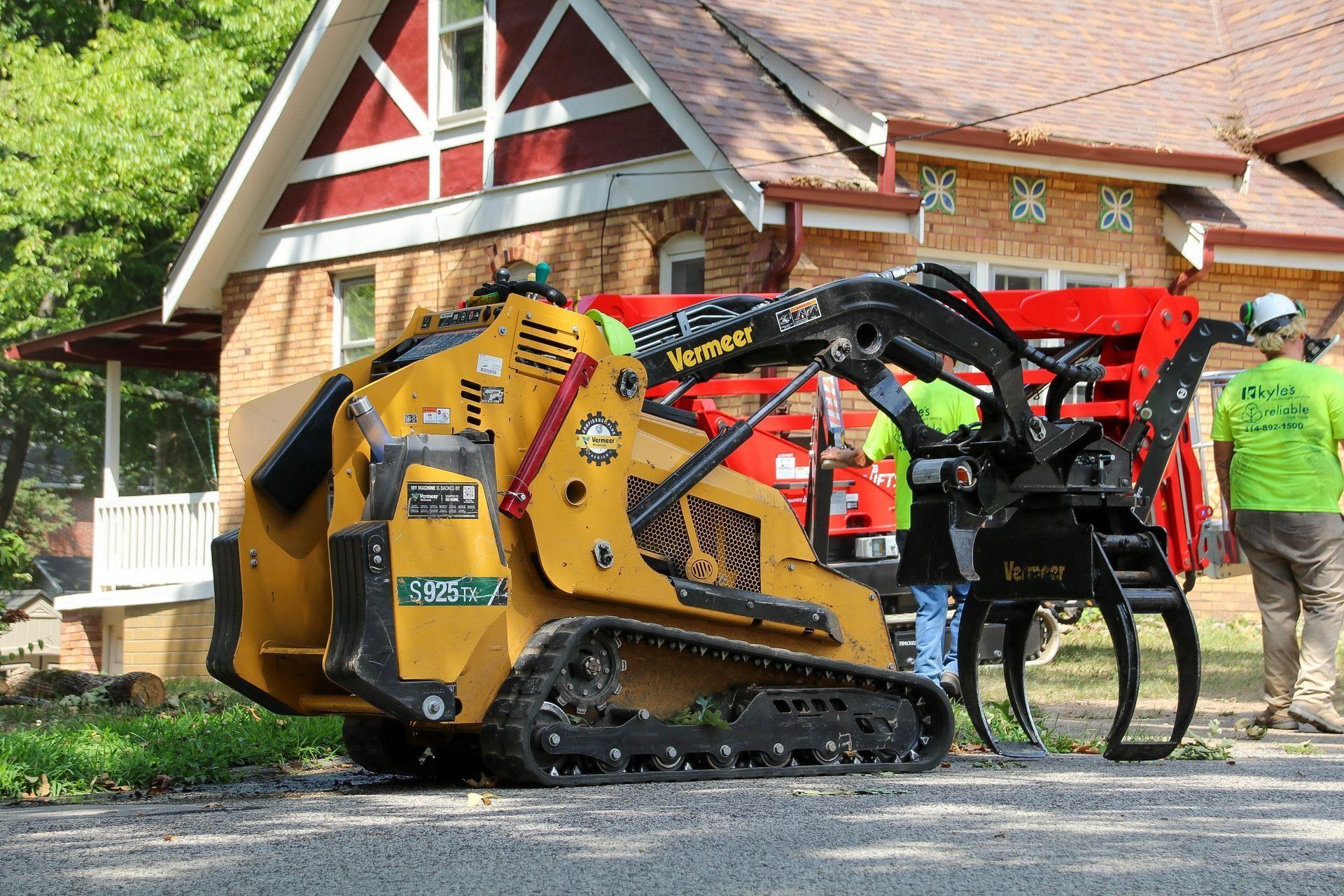 Yellow compact track loader with grapple attachment in front of a house, being operated by two people.