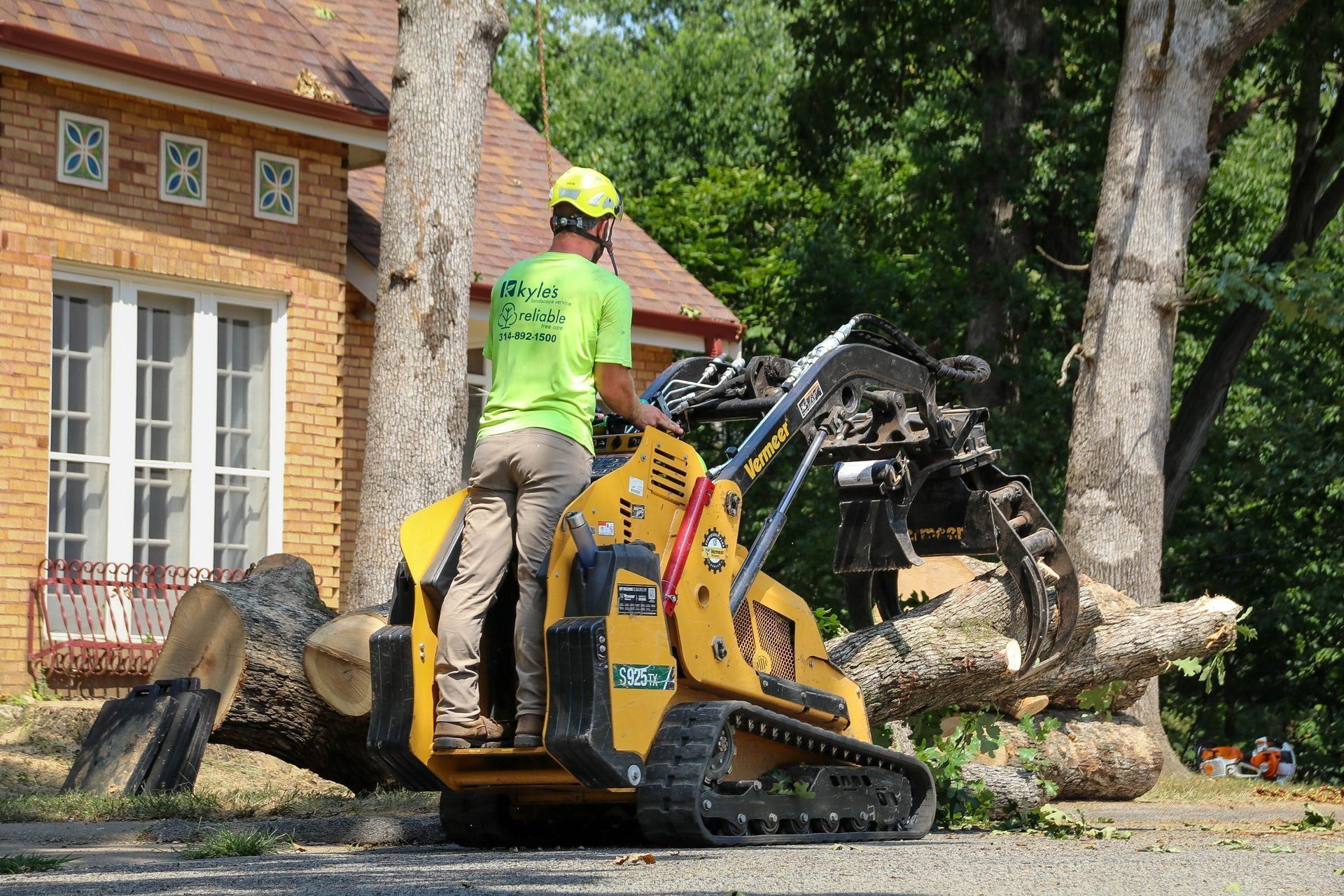 Man operating a yellow stump grinder next to a tree, near a brick house.