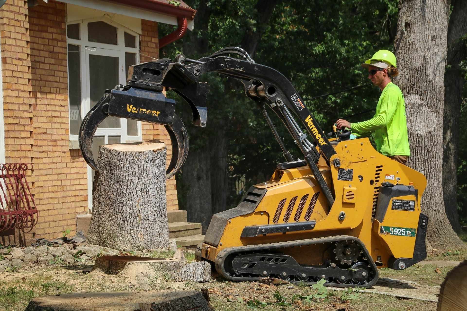 Yellow skid steer with grapple arm grinding a tree stump, operated by a person in a lime green shirt and hard hat.
