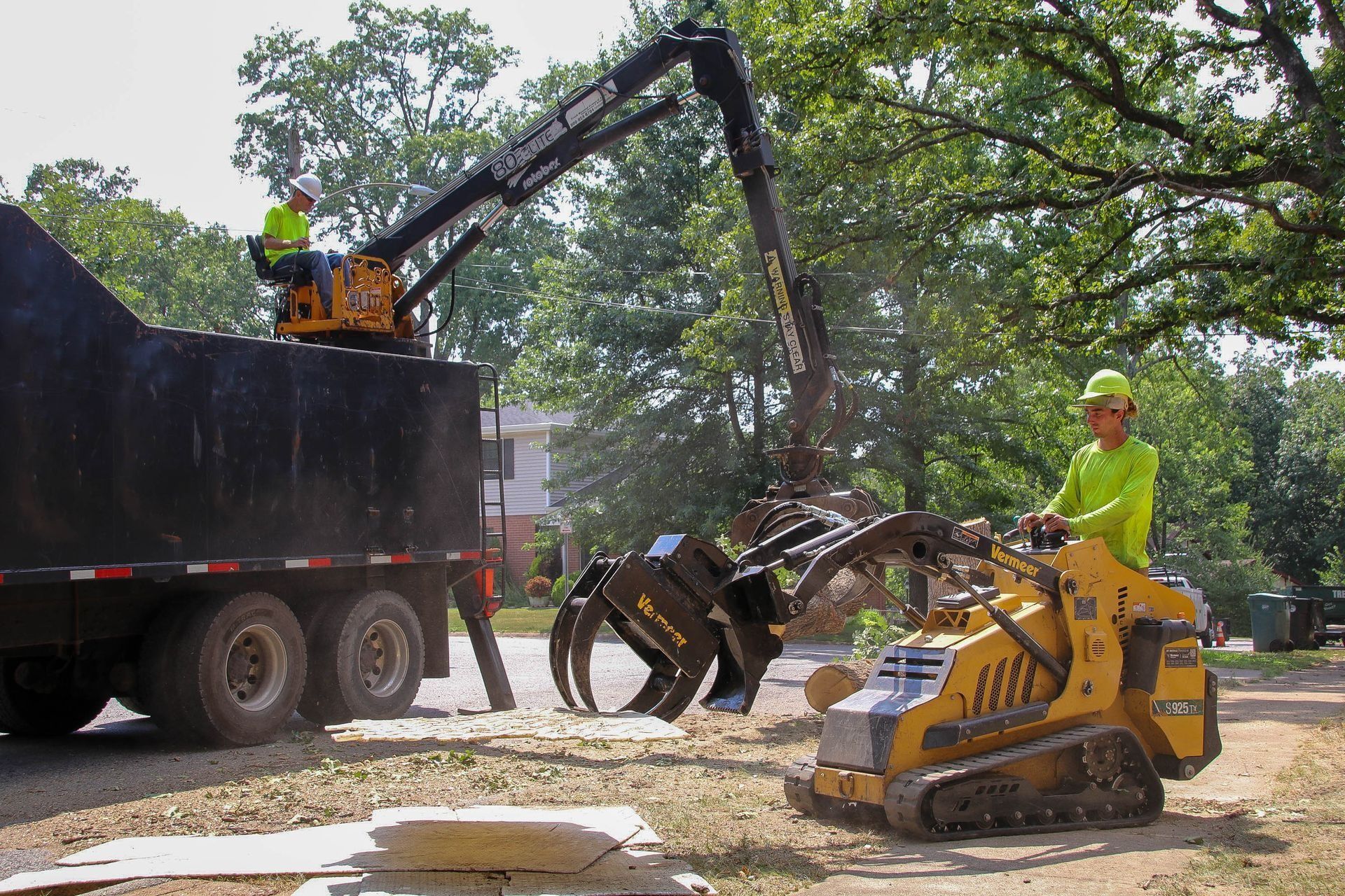 Two workers loading tree debris into a dump truck with an excavator and a skid steer in a residential area.