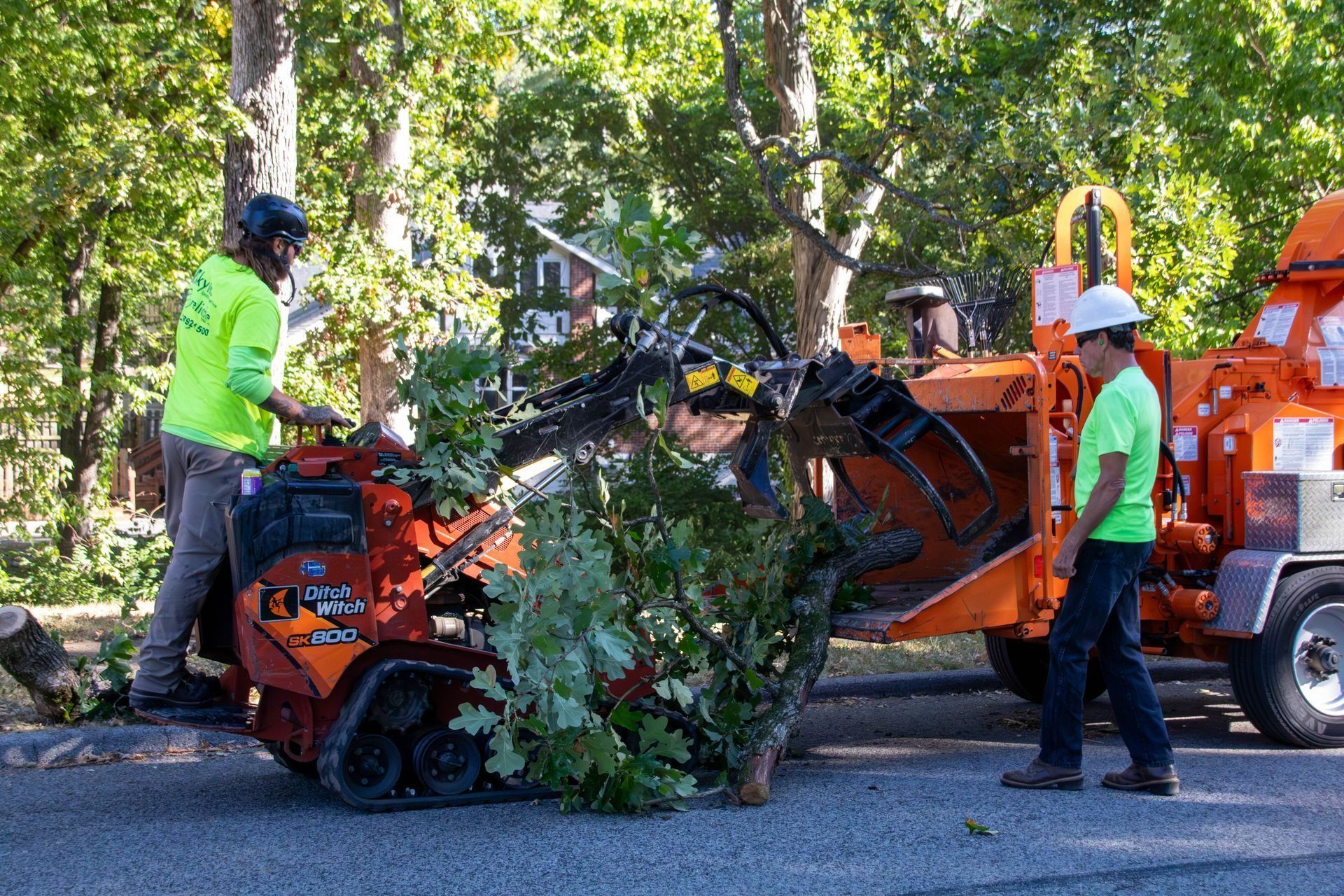 Two workers feeding tree branches into a wood chipper on a street. One operates a small tracked machine.
